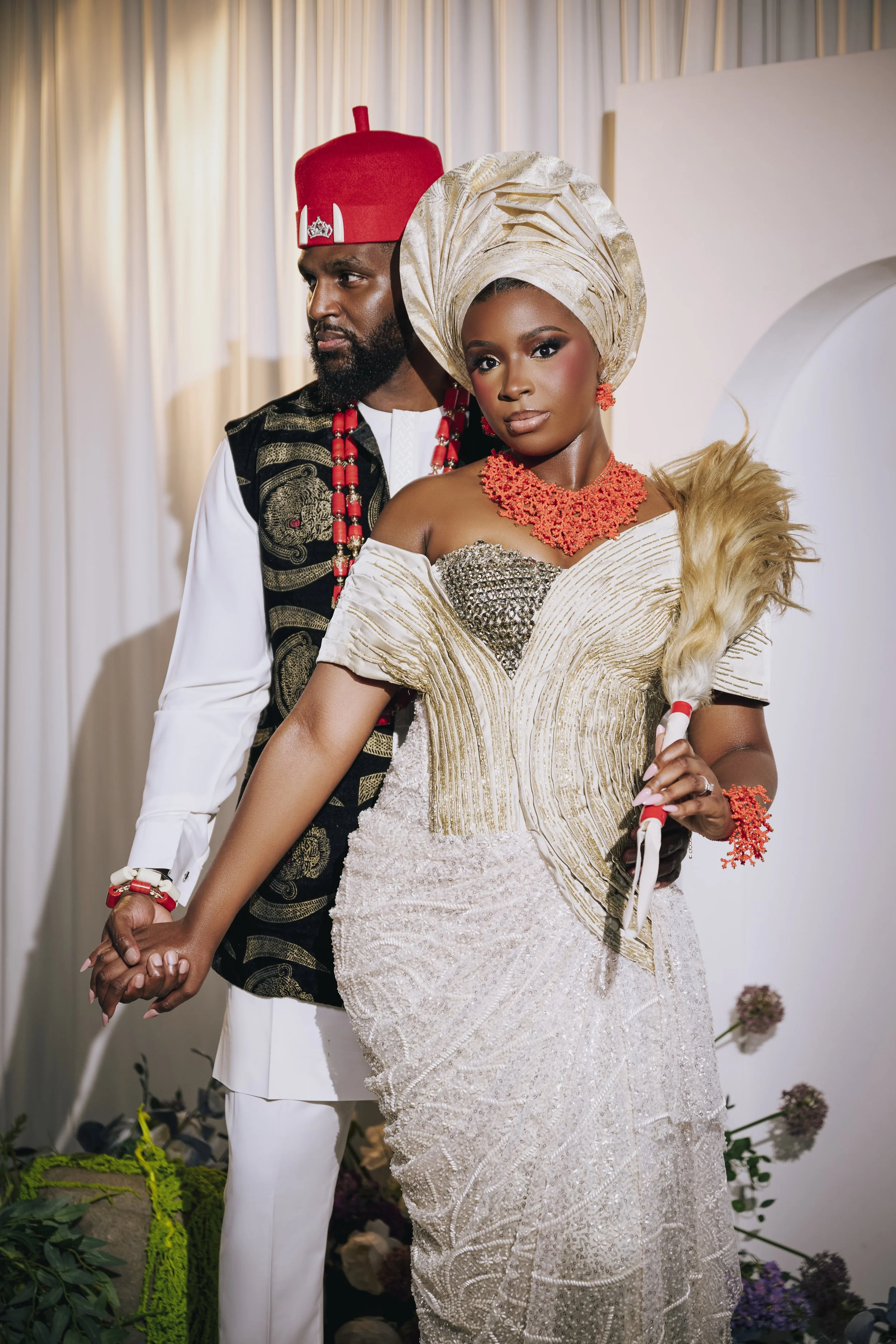 A couple in traditional African attire, holding hands, at a formal event. The woman is wearing an ornate, beige gown with matching headwrap and jewelry. The man is dressed in a white outfit with a black and gold vest and a red hat.