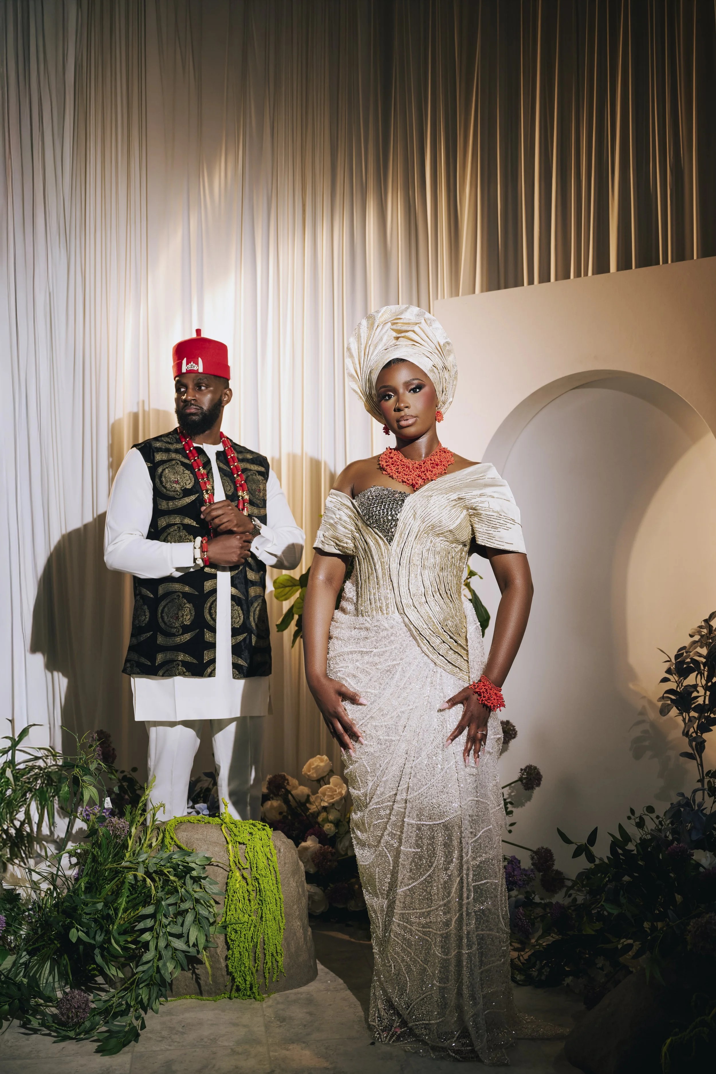 A man and woman dressed in traditional Nigerian attire, standing indoors with a backdrop of beige curtains and surrounding plants and flowers.