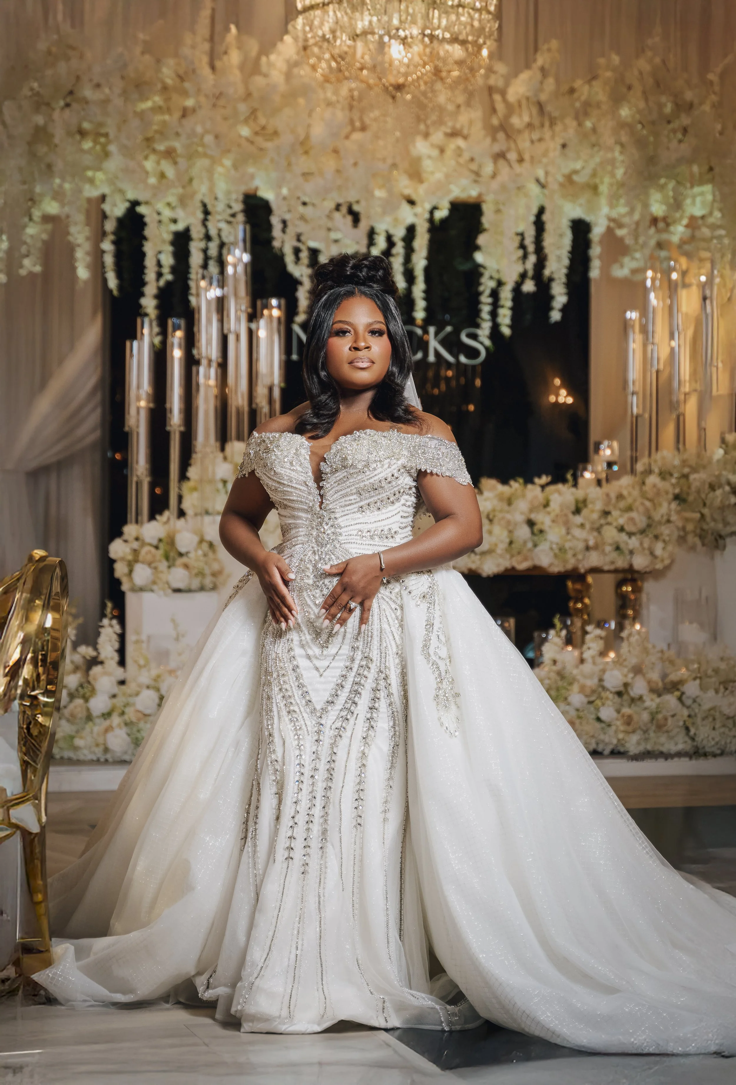 A woman in a white wedding dress with intricate embellishments, standing in front of a floral backdrop with candles and a chandelier.