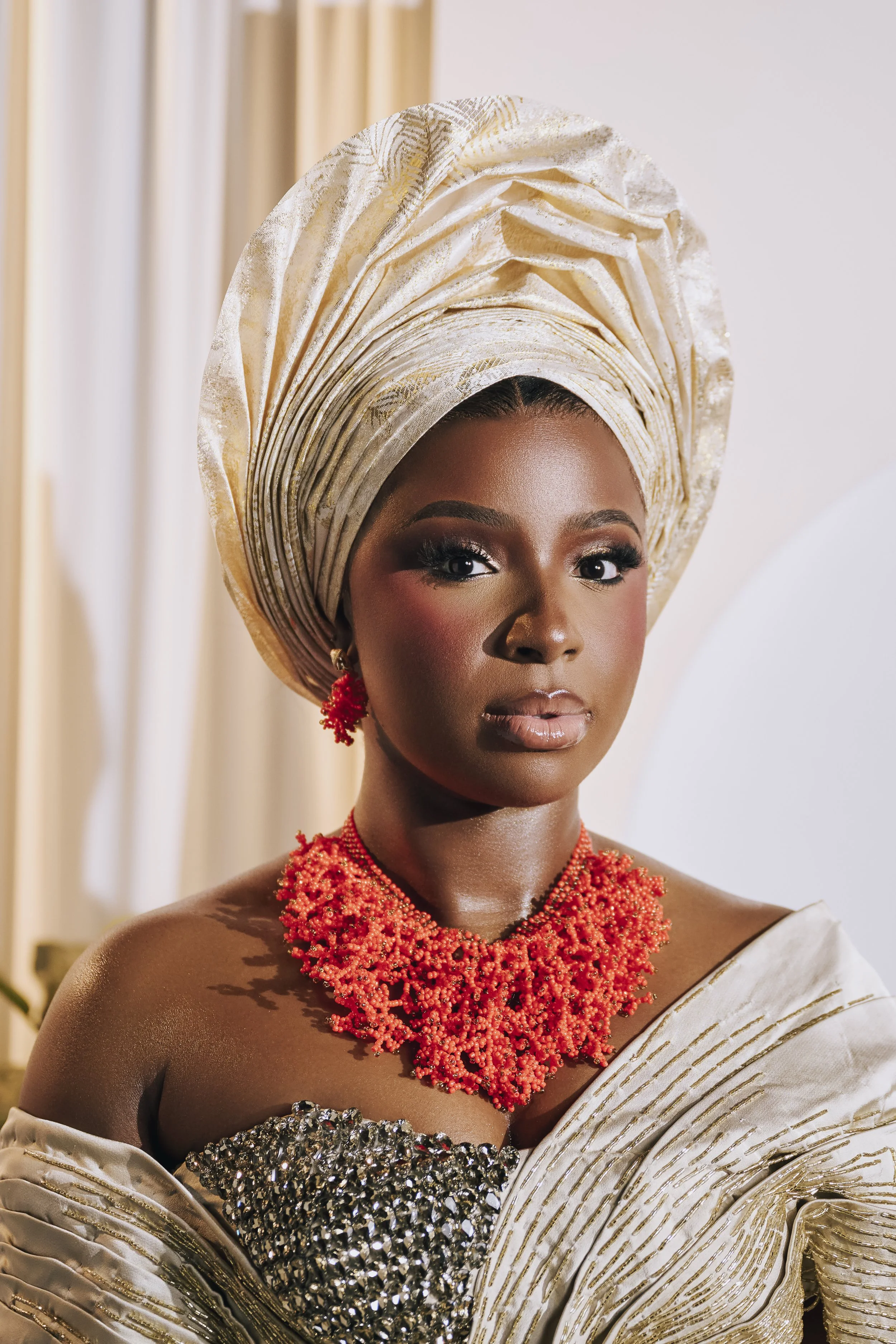 A woman dressed in traditional African attire, wearing a golden headwrap, a beaded red necklace, and a matching red earring, with elaborate makeup and a confident expression.