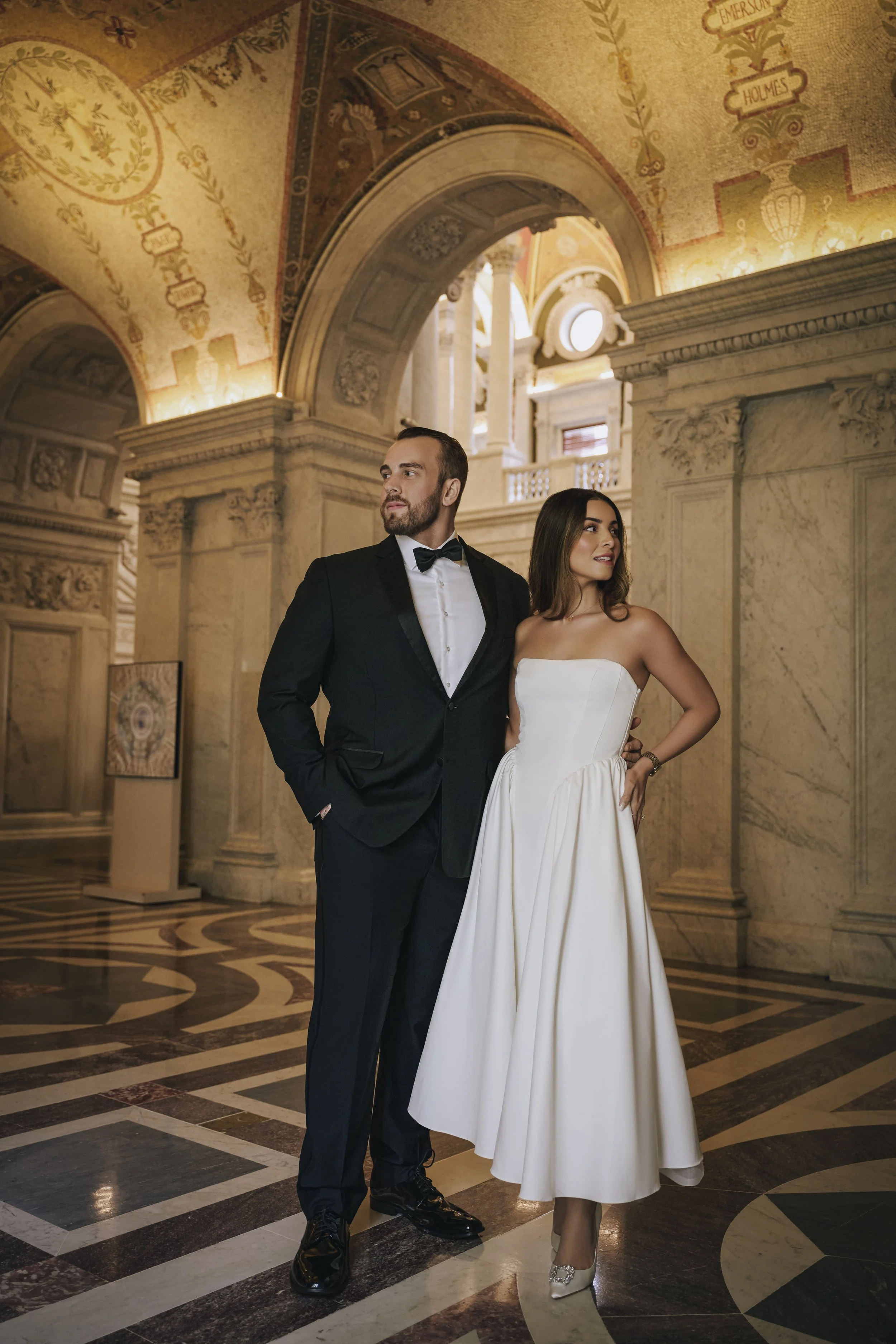 A man in a black tuxedo with a bow tie and a woman in a strapless white wedding dress and sparkling heels stand in a grand, ornately decorated indoor setting with columns and detailed ceiling art.