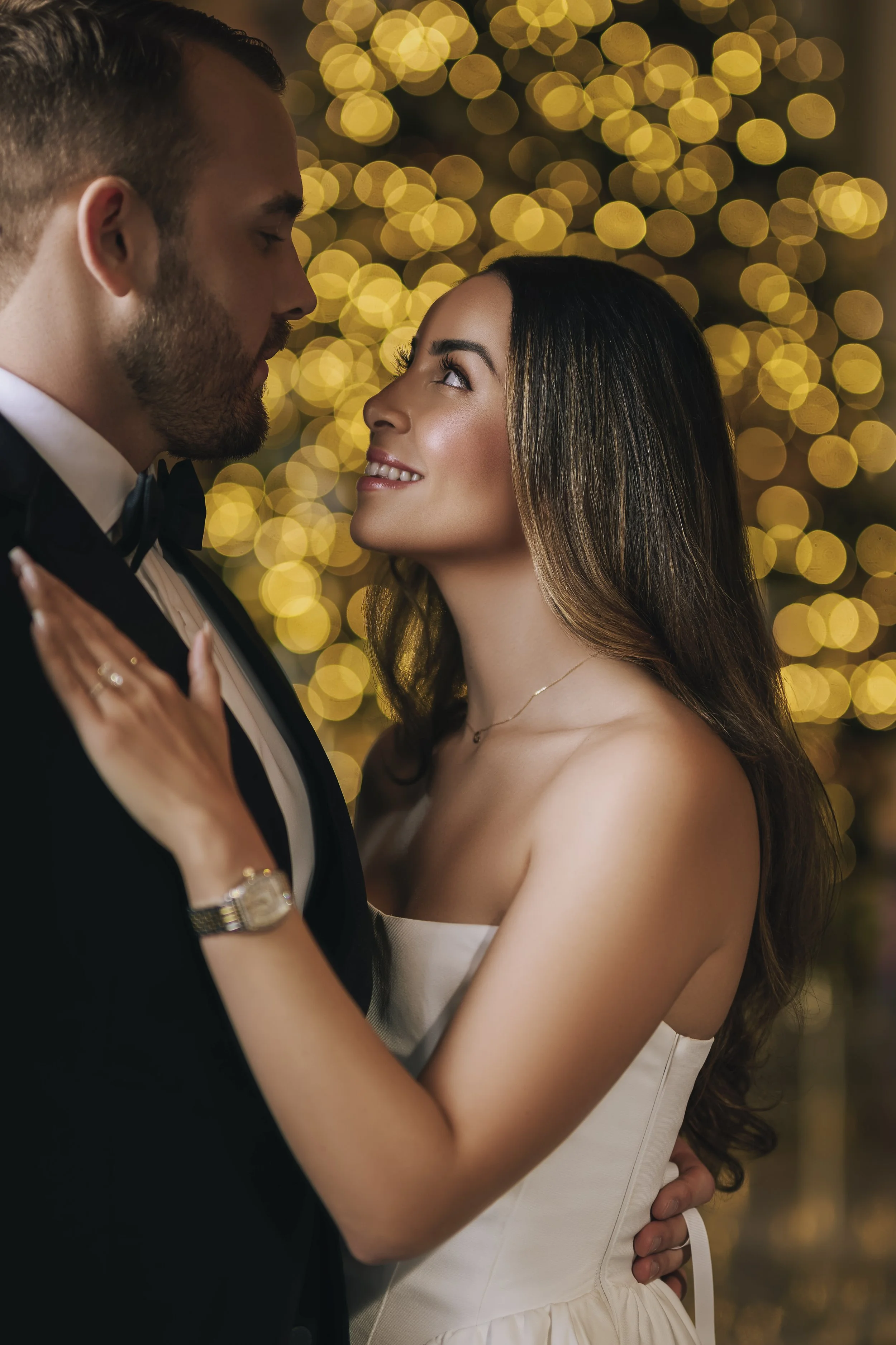 A man and woman in formal attire, standing close and gazing into each other's eyes, with a blurred background of warm string lights, creating a romantic ambience.