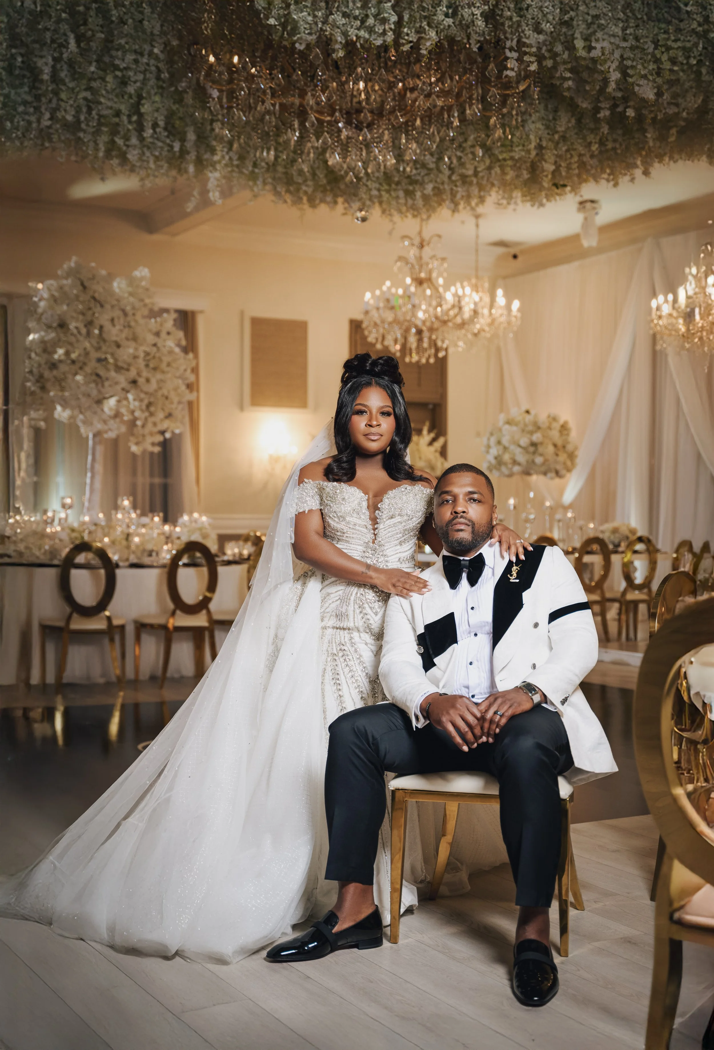 A bride in a white wedding dress and a groom in a tuxedo pose inside a wedding reception hall decorated with chandeliers, flowers, and elegant drapery. The bride is standing behind the seated groom, resting her hand on his shoulder.