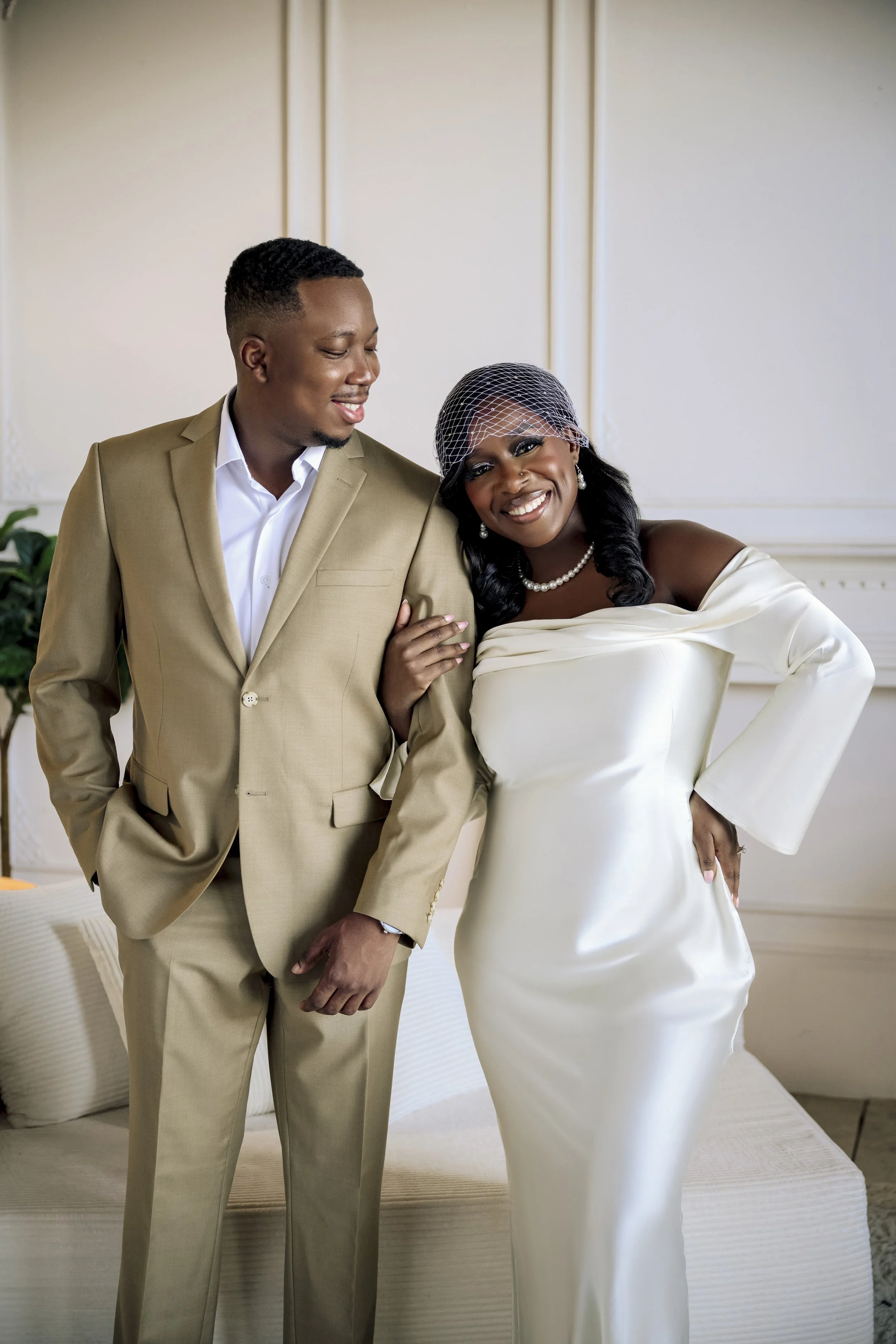 A smiling couple dressed in formal wedding attire standing together indoors, the woman wearing a white gown, pearl jewelry, and a birdcage veil, and the man in a beige suit with a white shirt.