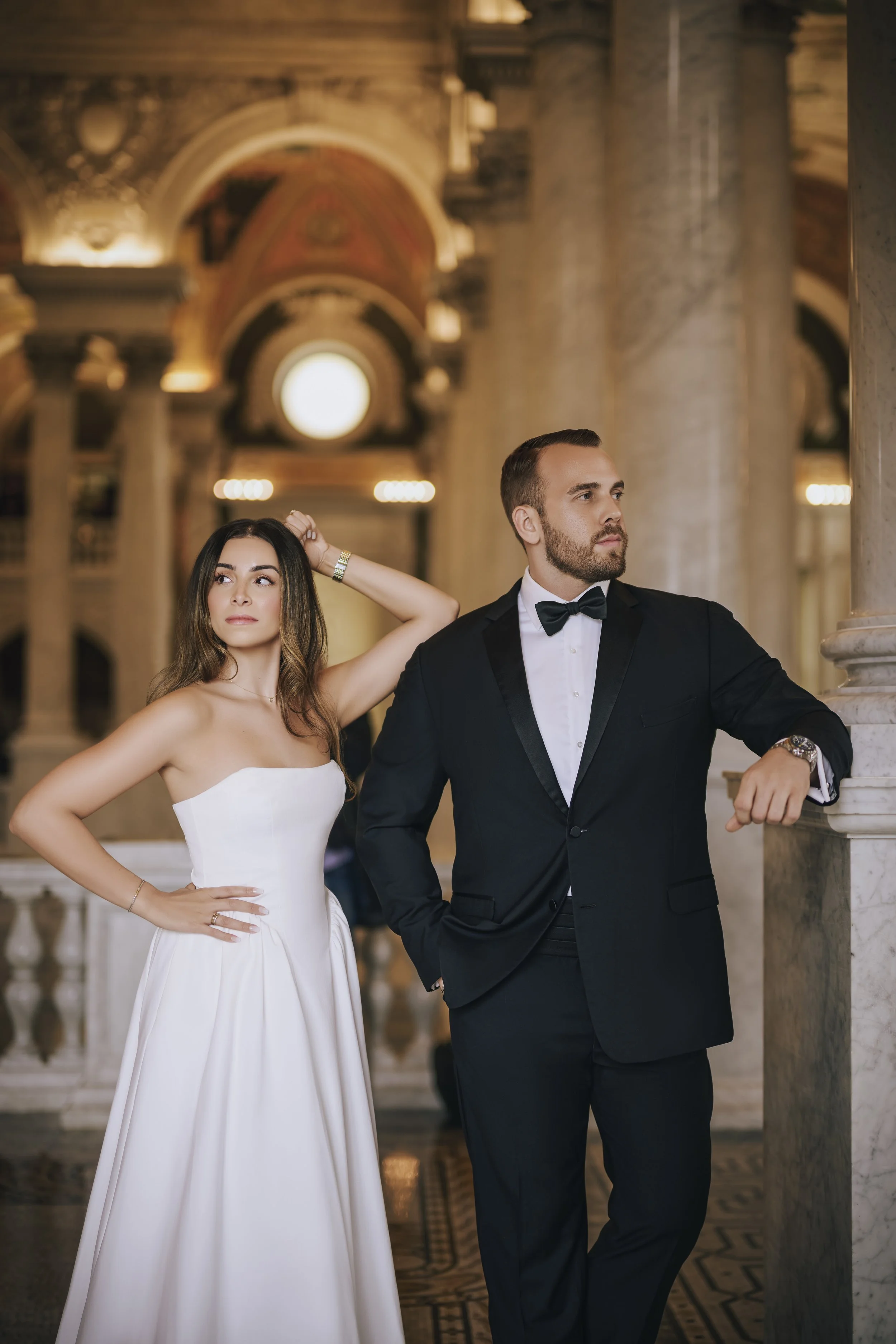 A woman in a white strapless dress and a man in a black tuxedo with a bow tie pose in an elegant, historic-looking building with ornate architecture and columns.