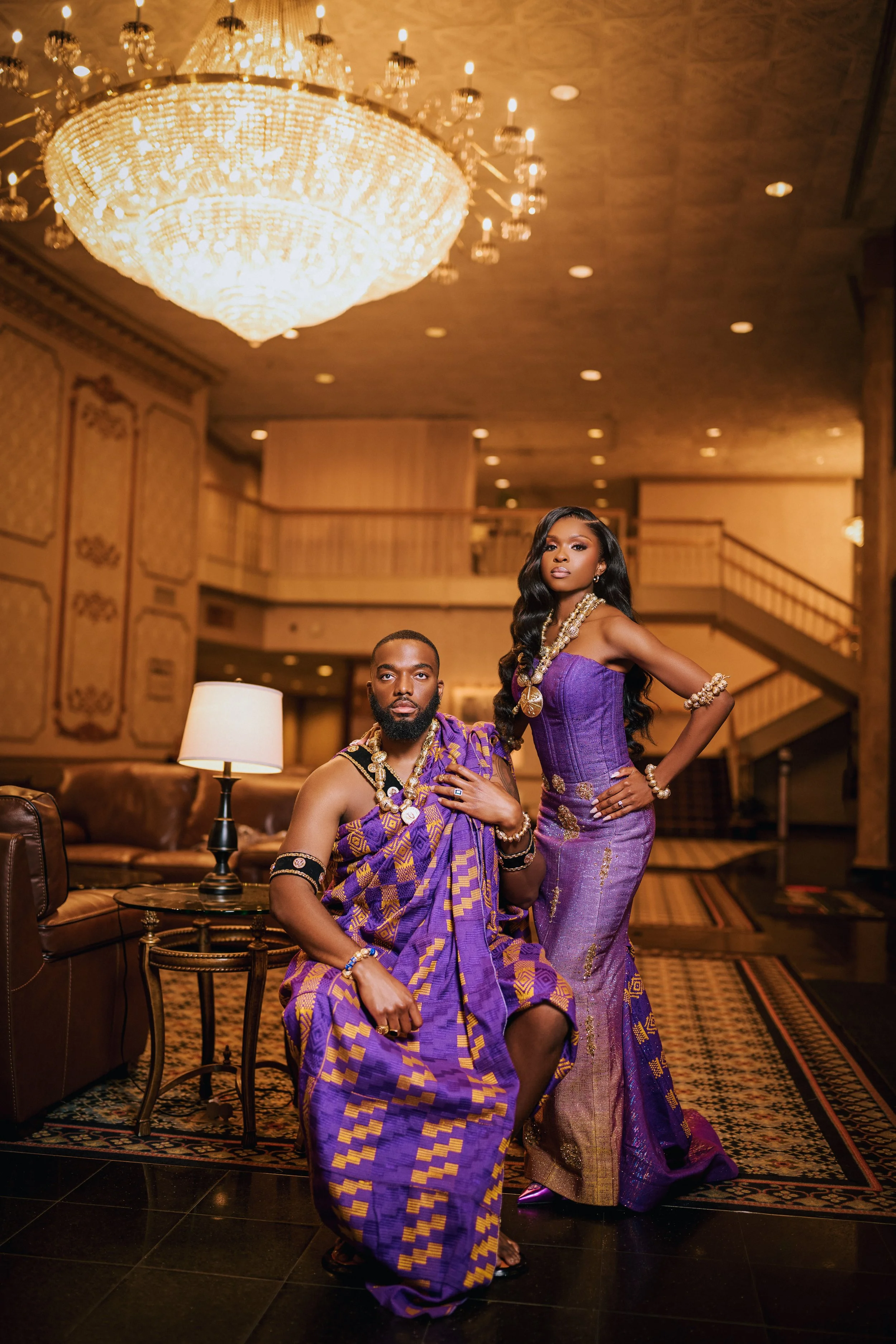 Two people of color dressed in purple traditional African attire with gold accents, jewelry, and accessories in a luxurious indoor setting with a chandelier and elegant decor.