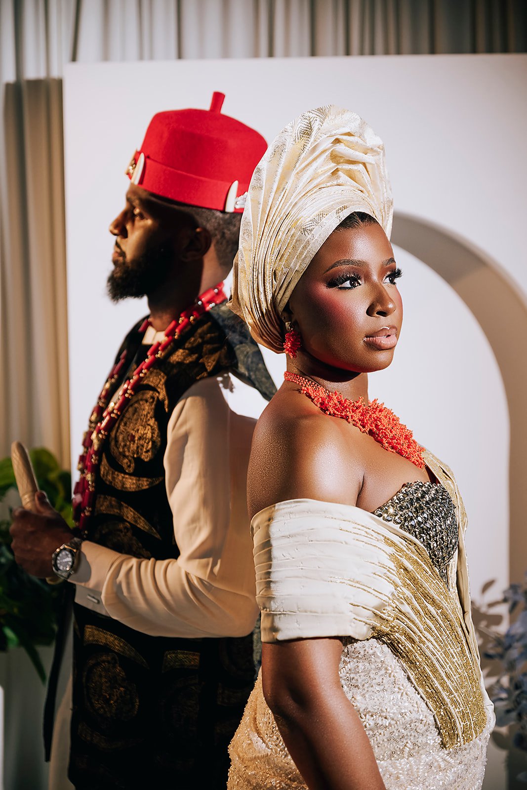 A woman in traditional African attire with a gold and white gele headwrap, coral jewelry, and a dress with gold and silver embellishments stands in front of a man dressed in traditional African clothing with a red hat, red beads, and a patterned garment, both posing indoors in front of a white backdrop.