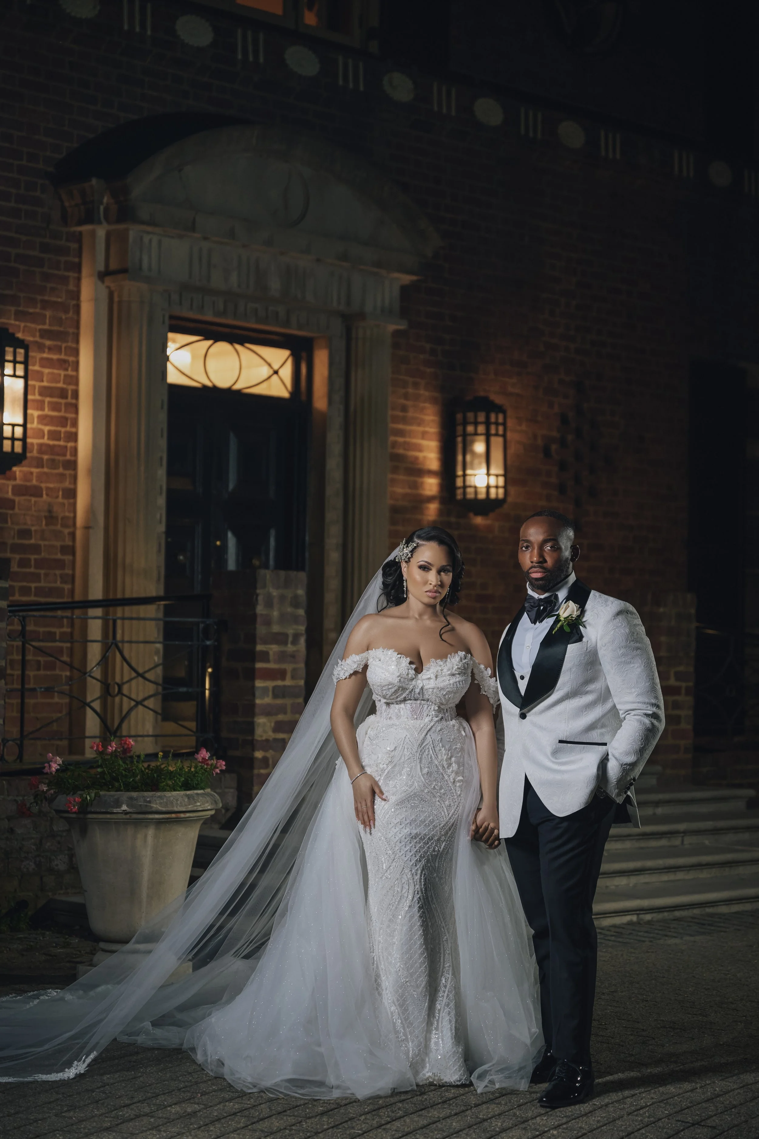 A bride and groom stand hand in hand outside a building at night, dressed in wedding attire. The bride wears a white off-the-shoulder wedding gown with a long veil, and the groom wears a white tuxedo with black lapels and a black bow tie. They are illuminated by warm lighting, with a brick building and outdoor lanterns in the background.