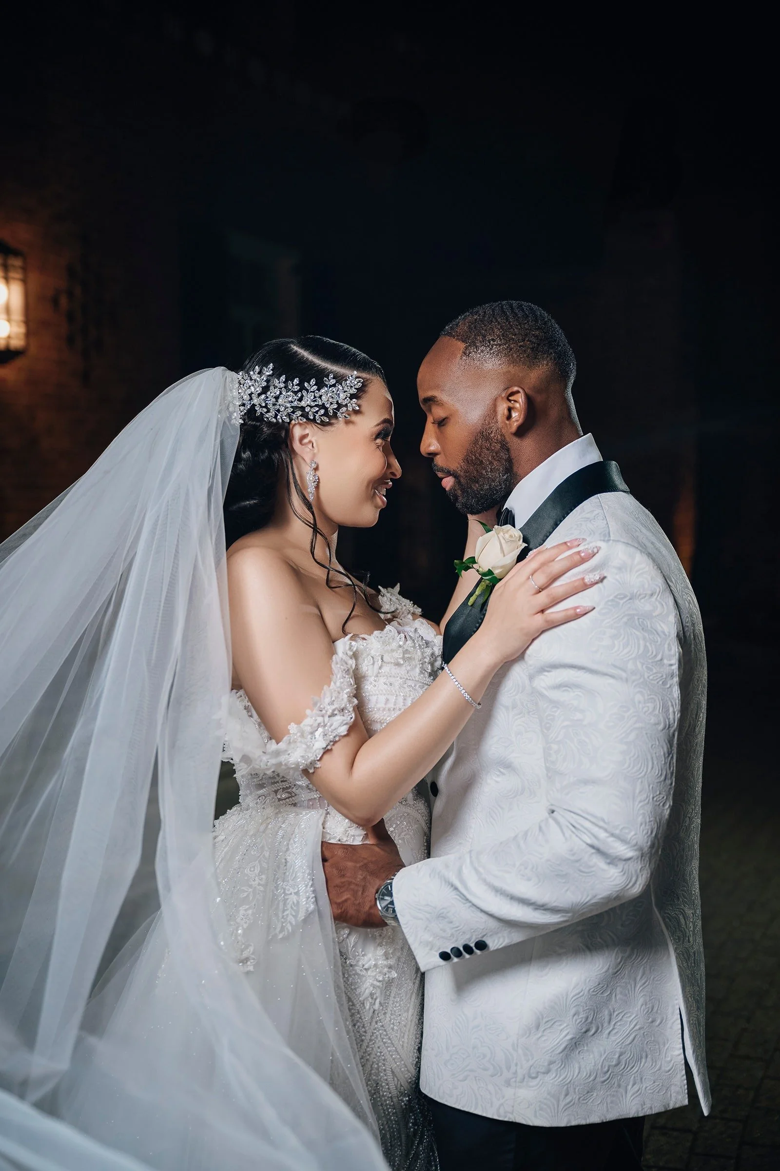 A bride and groom are facing each other closely, with their foreheads touching and eyes closed, at night with a dark background. The bride is wearing a white wedding dress with lace details, a veil, and floral headpiece. The groom is dressed in a white tuxedo with a textured pattern, black bow tie, and a white rose boutonniere.