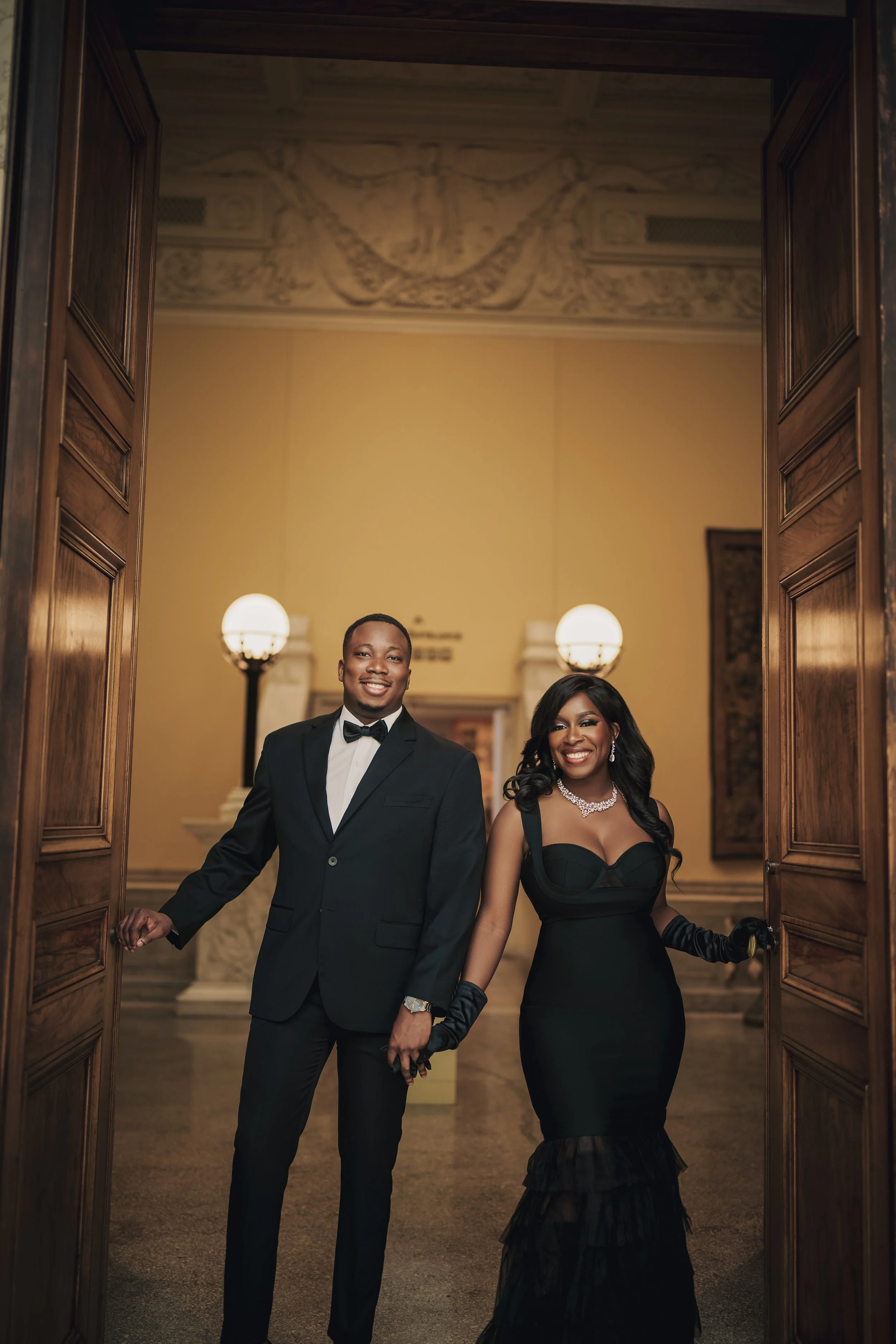 A smiling young man in a tuxedo and a woman in a black evening gown standing inside a grand building with wooden doors, warm lighting, and elegant decor.