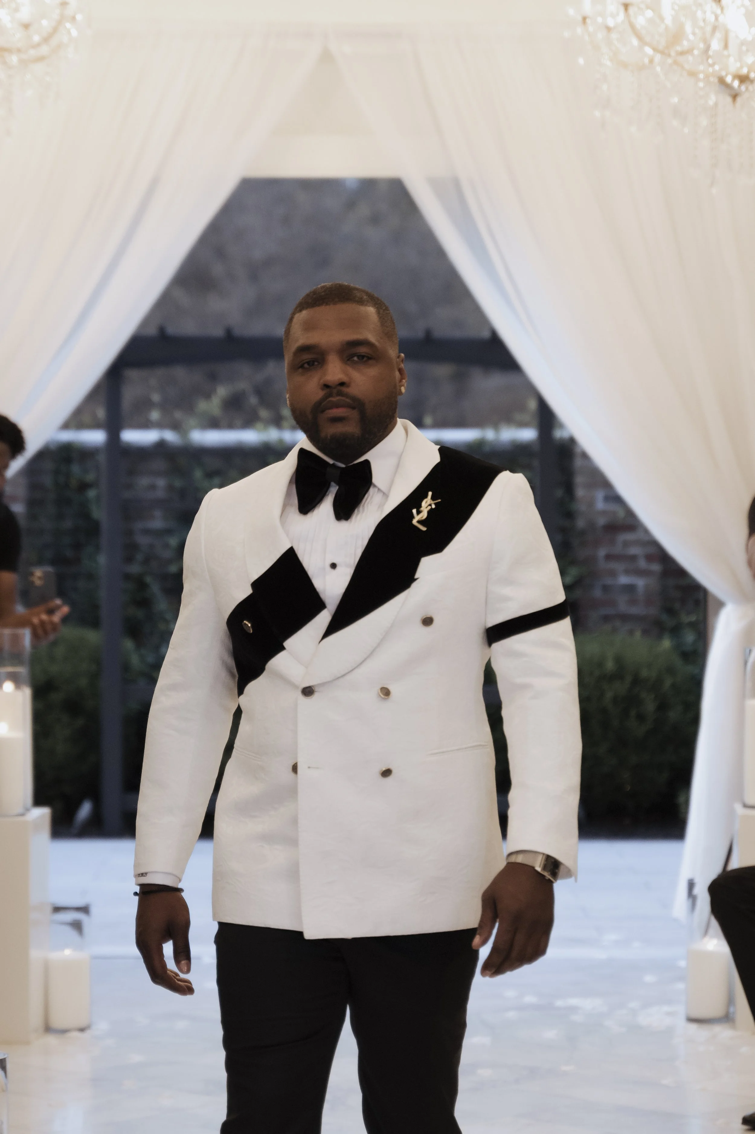 A man in a white tuxedo with black accents, wearing a black bow tie, walking in a decorated indoor space with draped fabric and chandeliers.