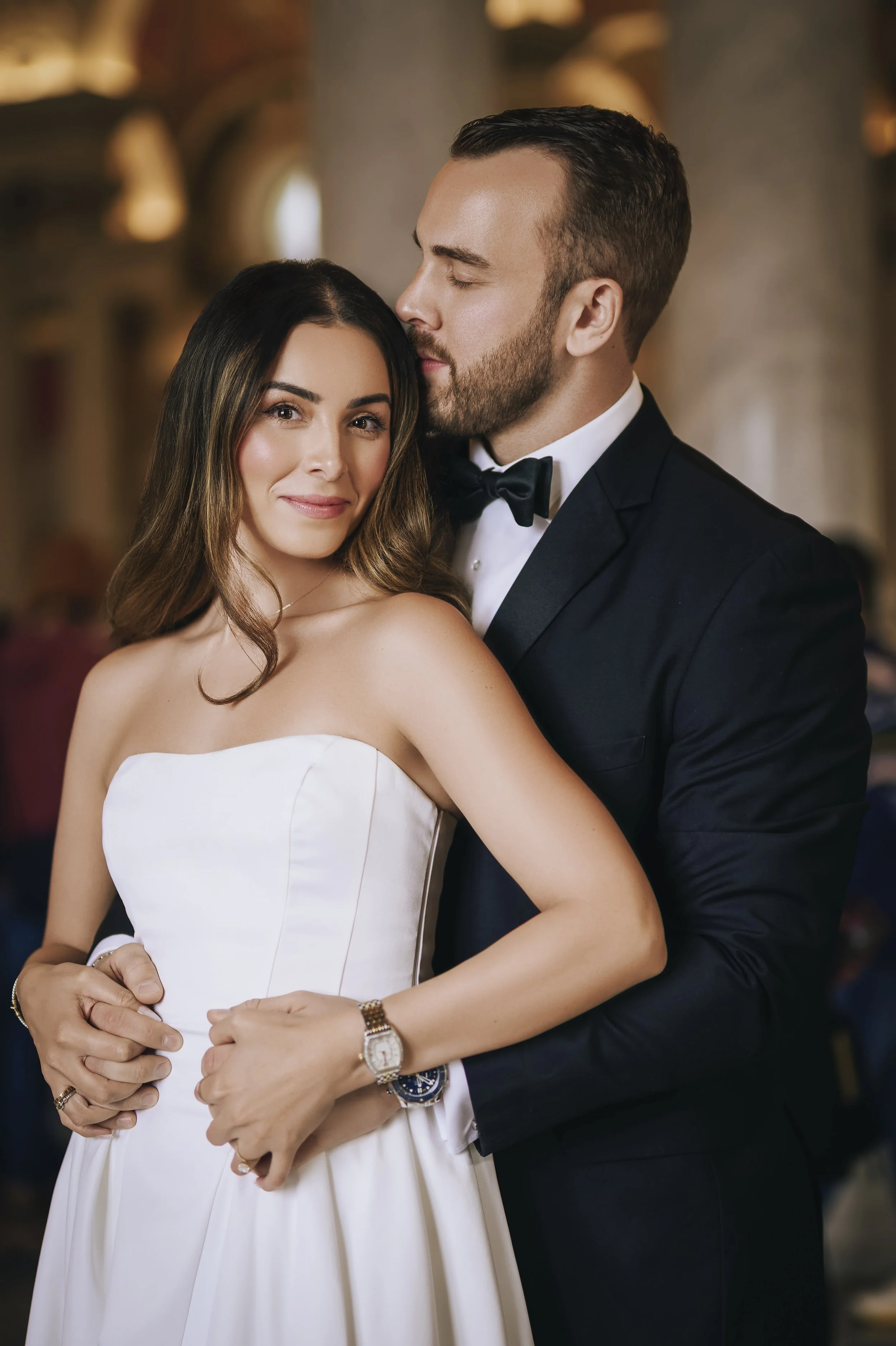 A couple in wedding attire, the woman in a white strapless wedding dress and the man in a black tuxedo, embracing affectionately at a wedding or formal event.