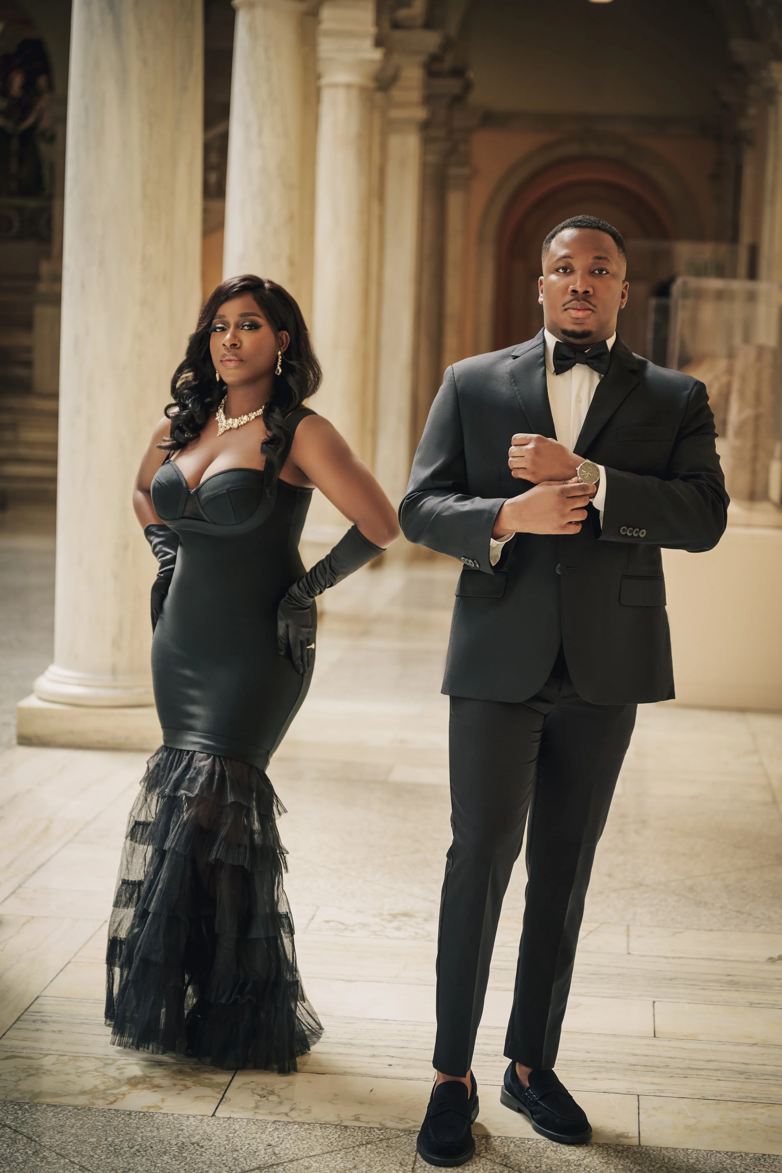 A man and woman dressed in formal attire standing in an elegant, grand hall with tall columns and ornate architectural details.