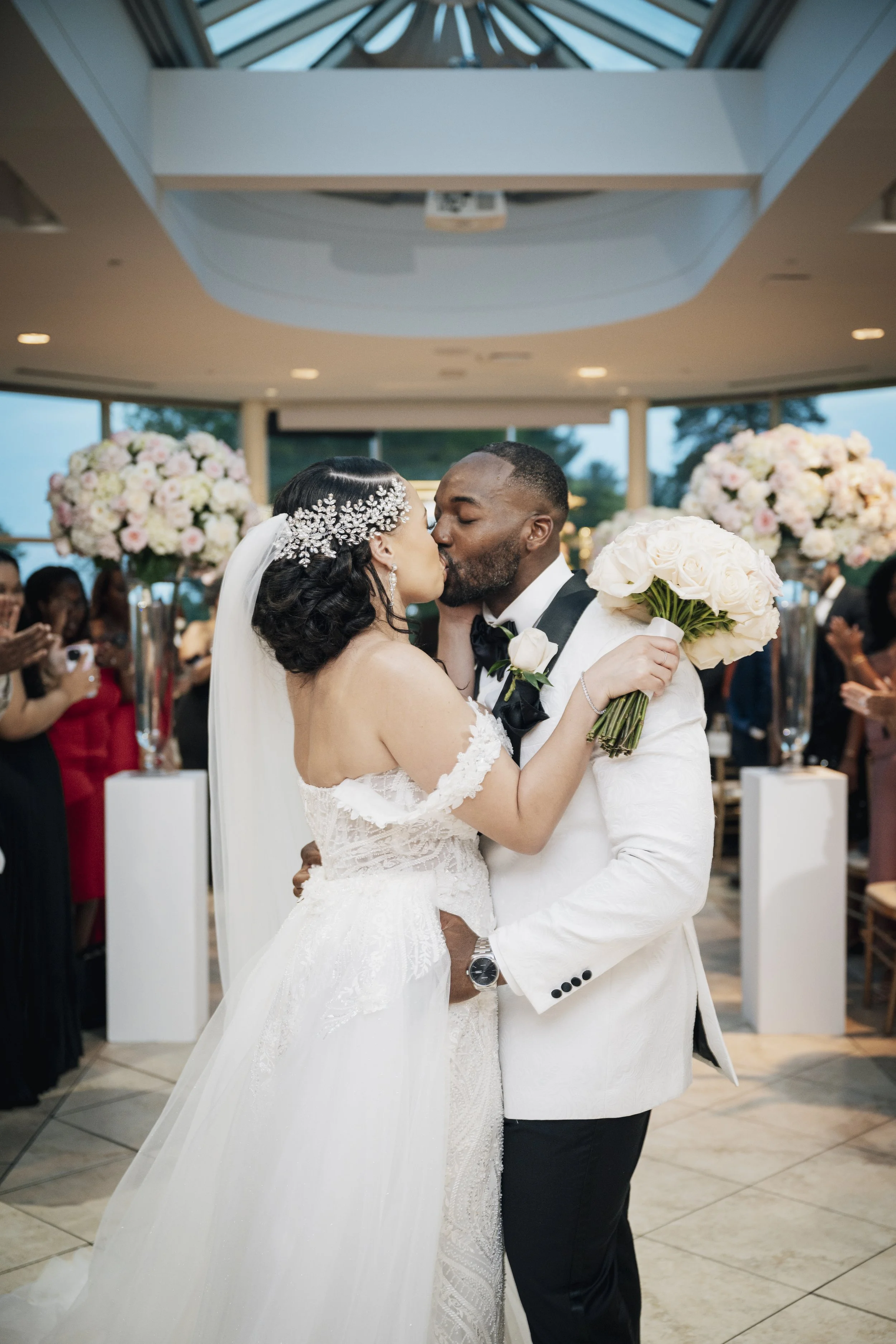 A bride and groom share a kiss during their wedding ceremony, with guests in the background and floral arrangements.