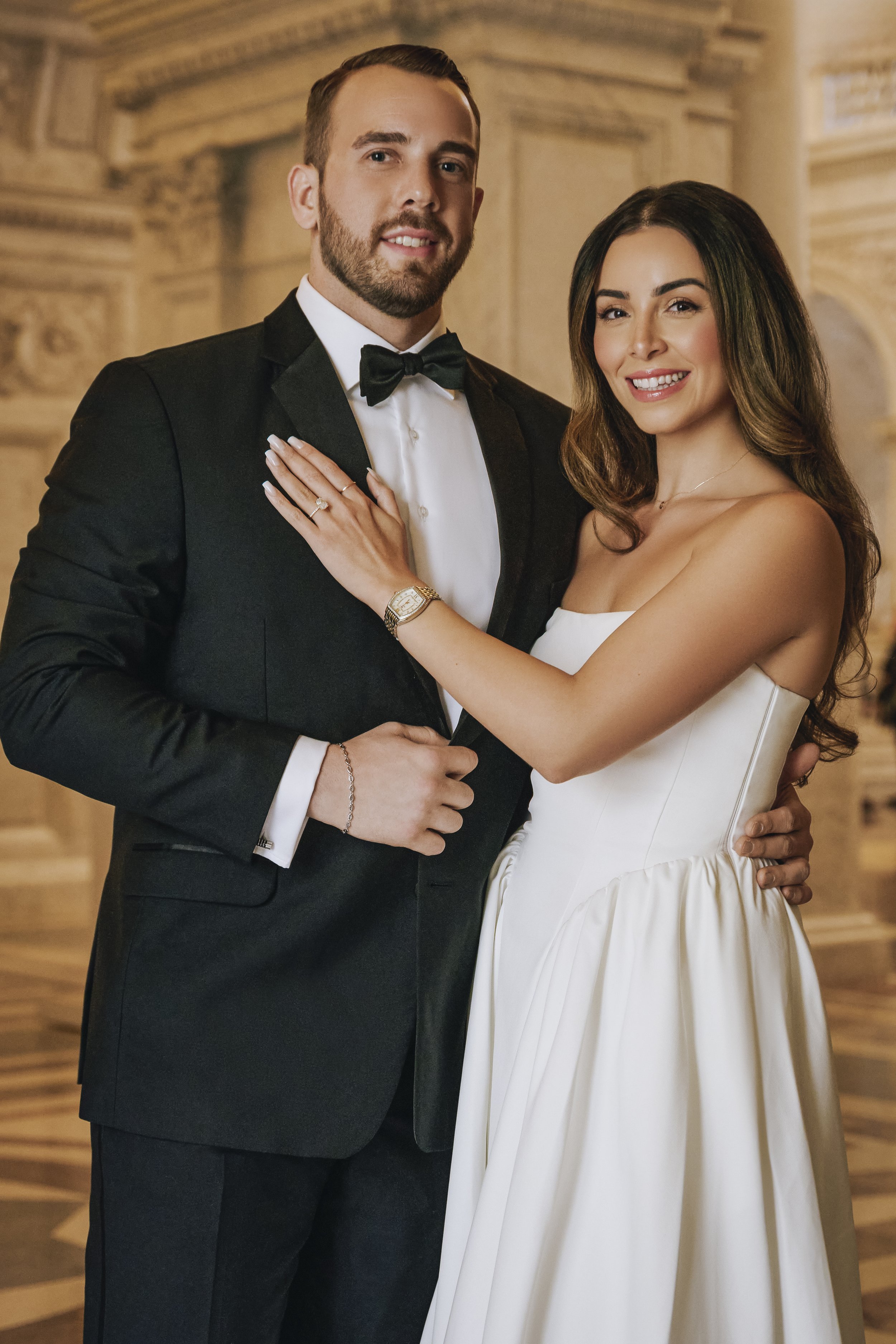 A newlywed couple posing indoors, the groom in a black tuxedo and bow tie, the bride in a white wedding dress, both smiling and showing their rings.