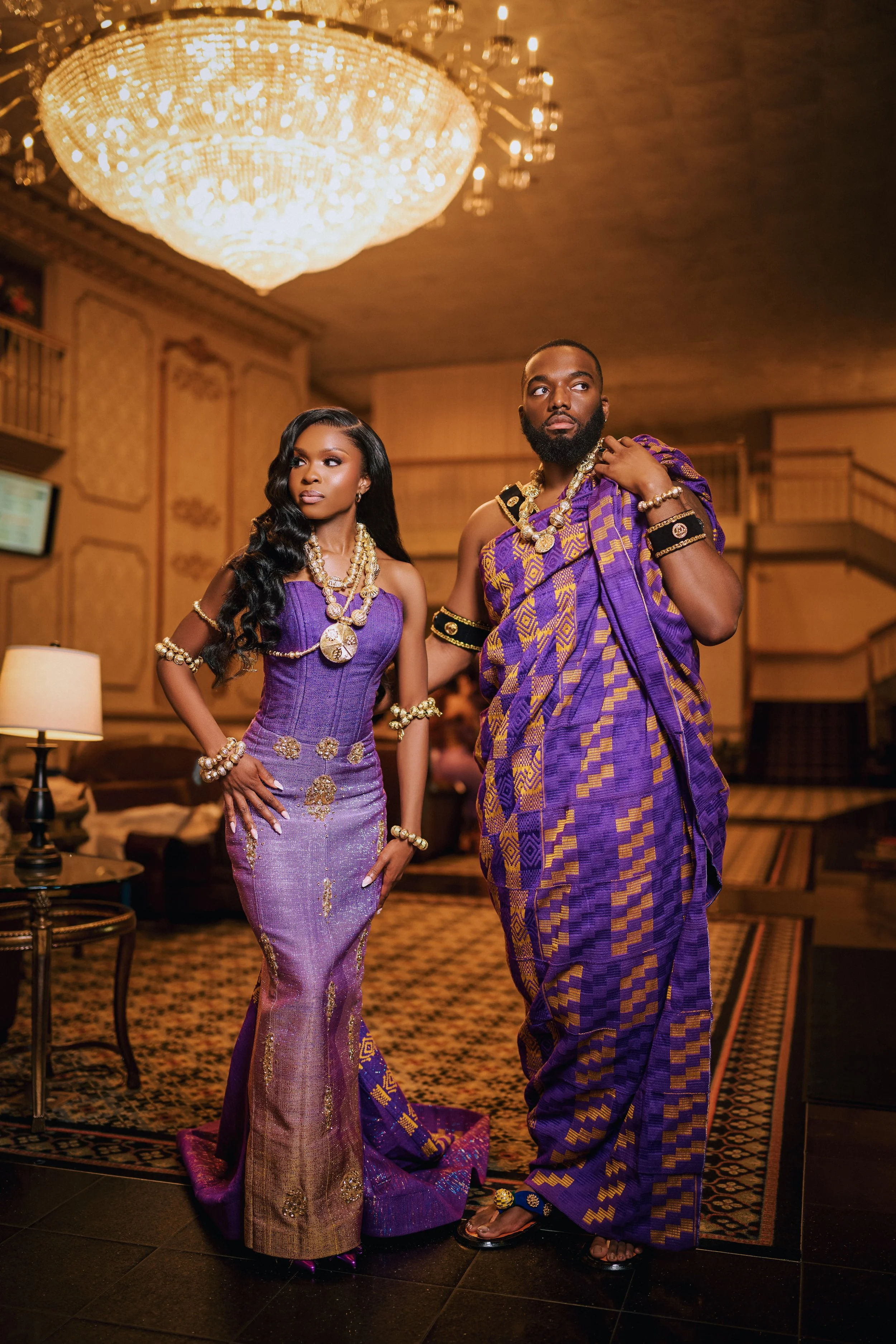 Young woman and man dressed in purple and gold traditional African attire, standing in a luxurious room with chandelier and ornate decor.