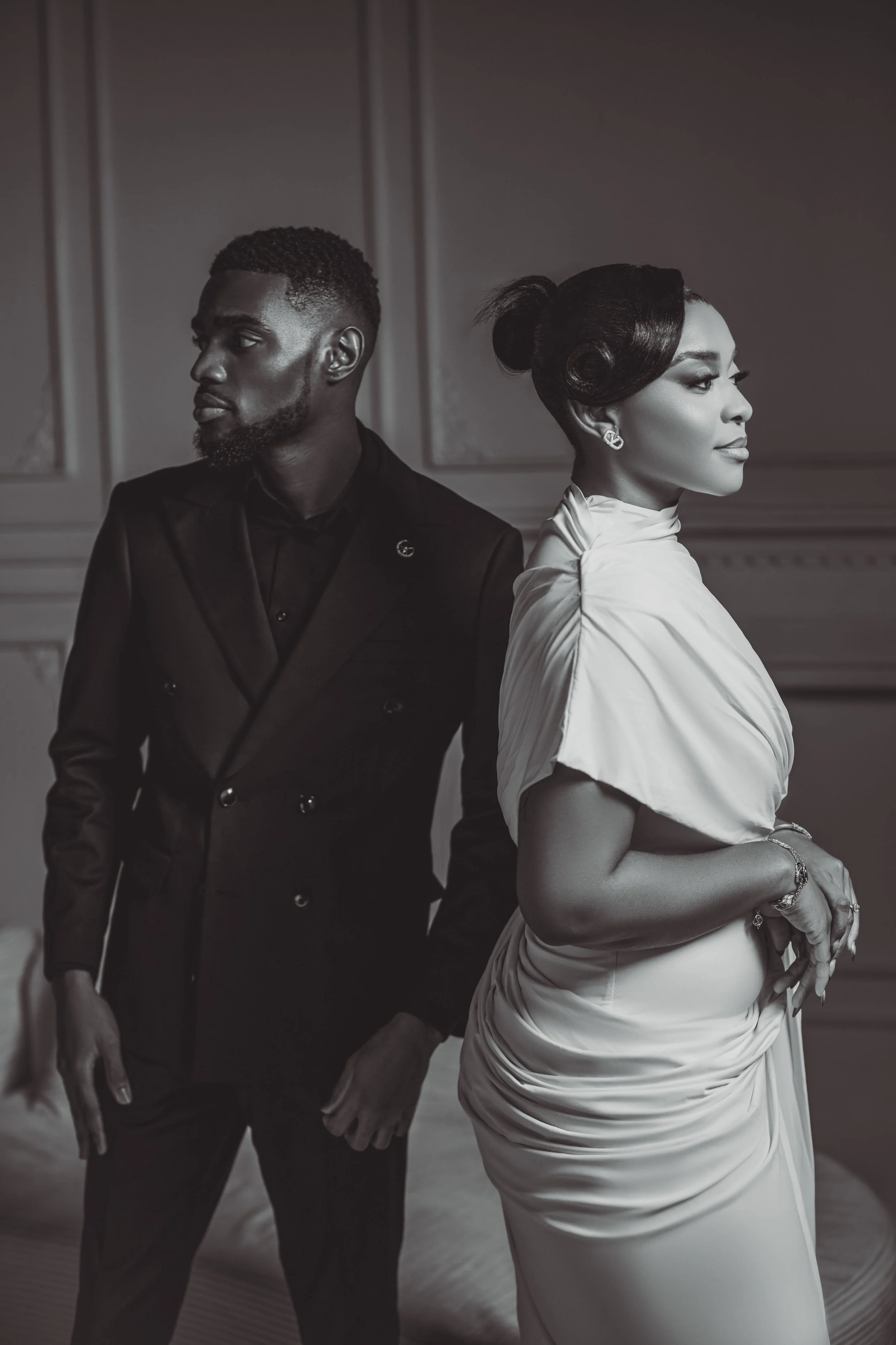 A stylishly dressed man and woman stand back-to-back in a formal setting with elegant wall paneling. The man wears a dark double-breasted suit and has a beard and short hair. The woman wears a sophisticated light-colored dress with ruffled sleeves, bay leaf earrings, and has an intricate vintage-inspired hairstyle.