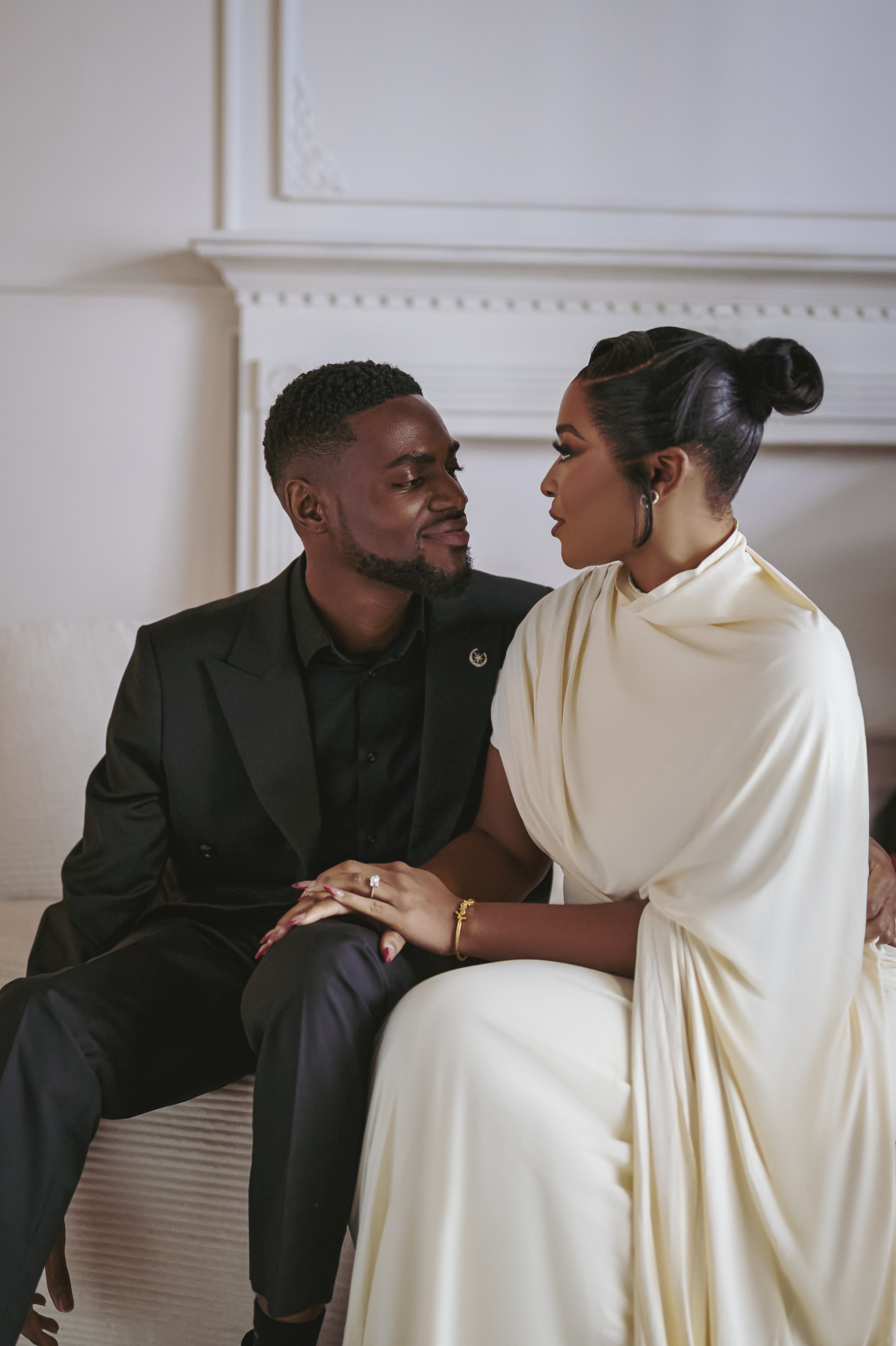 A couple sitting close together, gazing into each other's eyes. The man is wearing a black suit and the woman a white dress, with her hair styled in an elegant updo. They are holding hands affectionately in a warmly lit room.