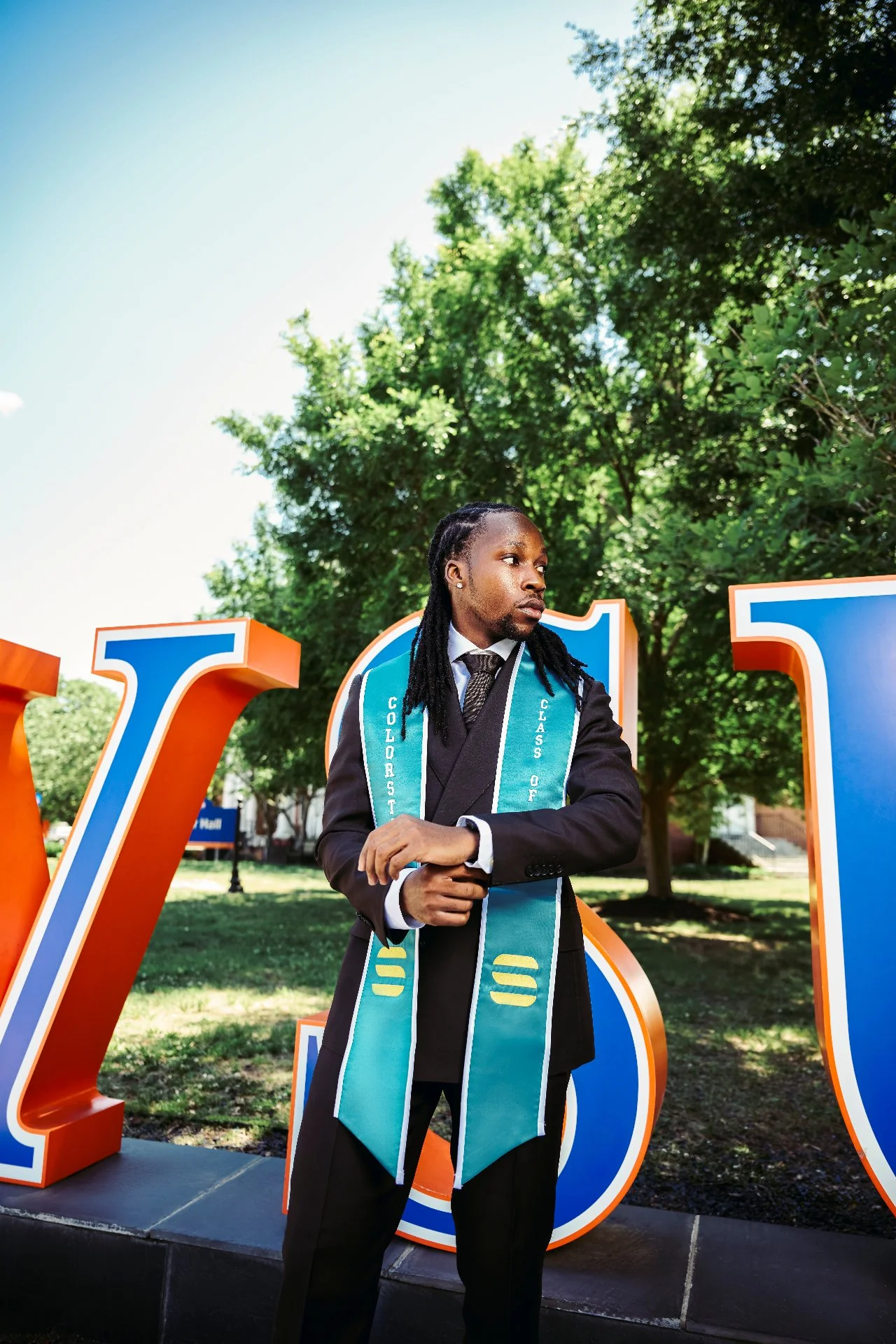 A man dressed in a suit and tie wearing a graduation stole, standing outdoors in front of large orange and blue letters, with trees and a park setting in the background.