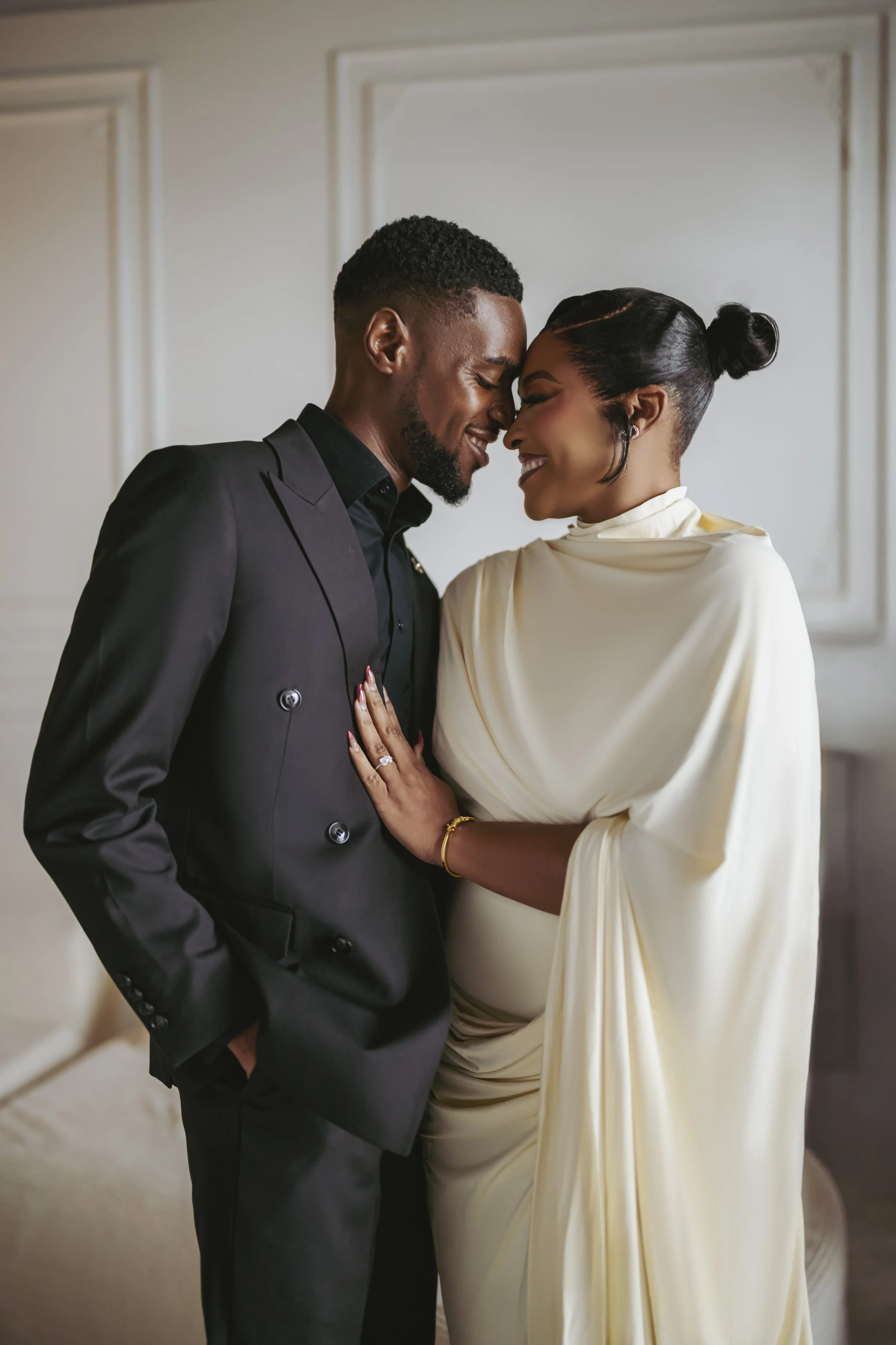 A couple is standing close, smiling and touching foreheads. The man is wearing a black suit with a shirt, and the woman is in a cream-colored satin dress with a high neck. They appear happy and intimate, in a neutral, softly lit indoor setting.