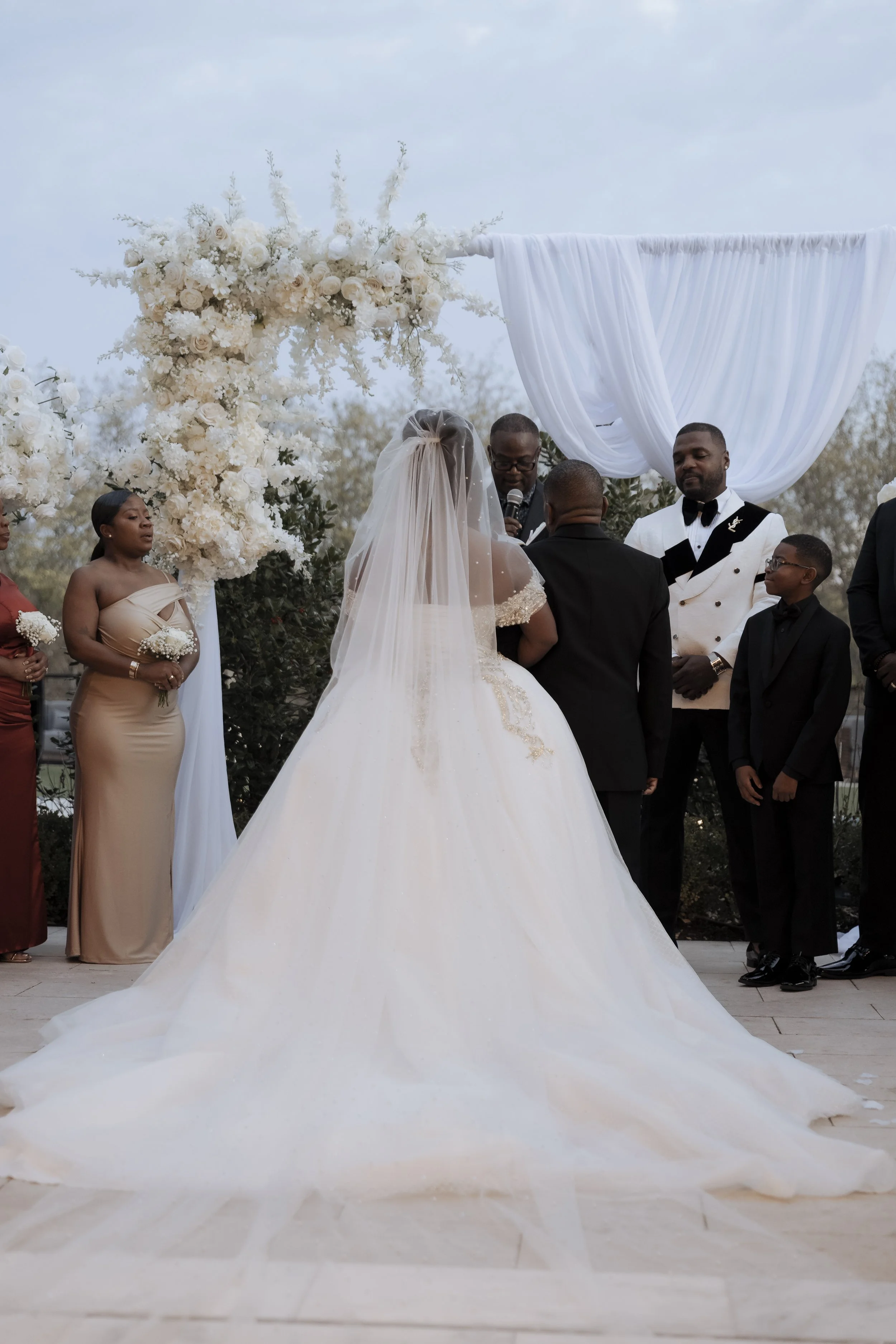 Wedding ceremony with bride and groom facing each other, officiant with microphone, wedding party members standing nearby, large white floral arrangement and white draped fabric backdrop.