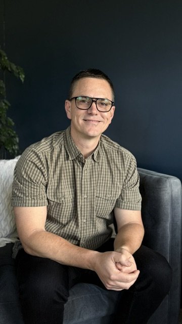A man in glasses and a checkered shirt sitting on a gray couch, smiling at the camera, with a dark blue wall and part of a plant in the background.
