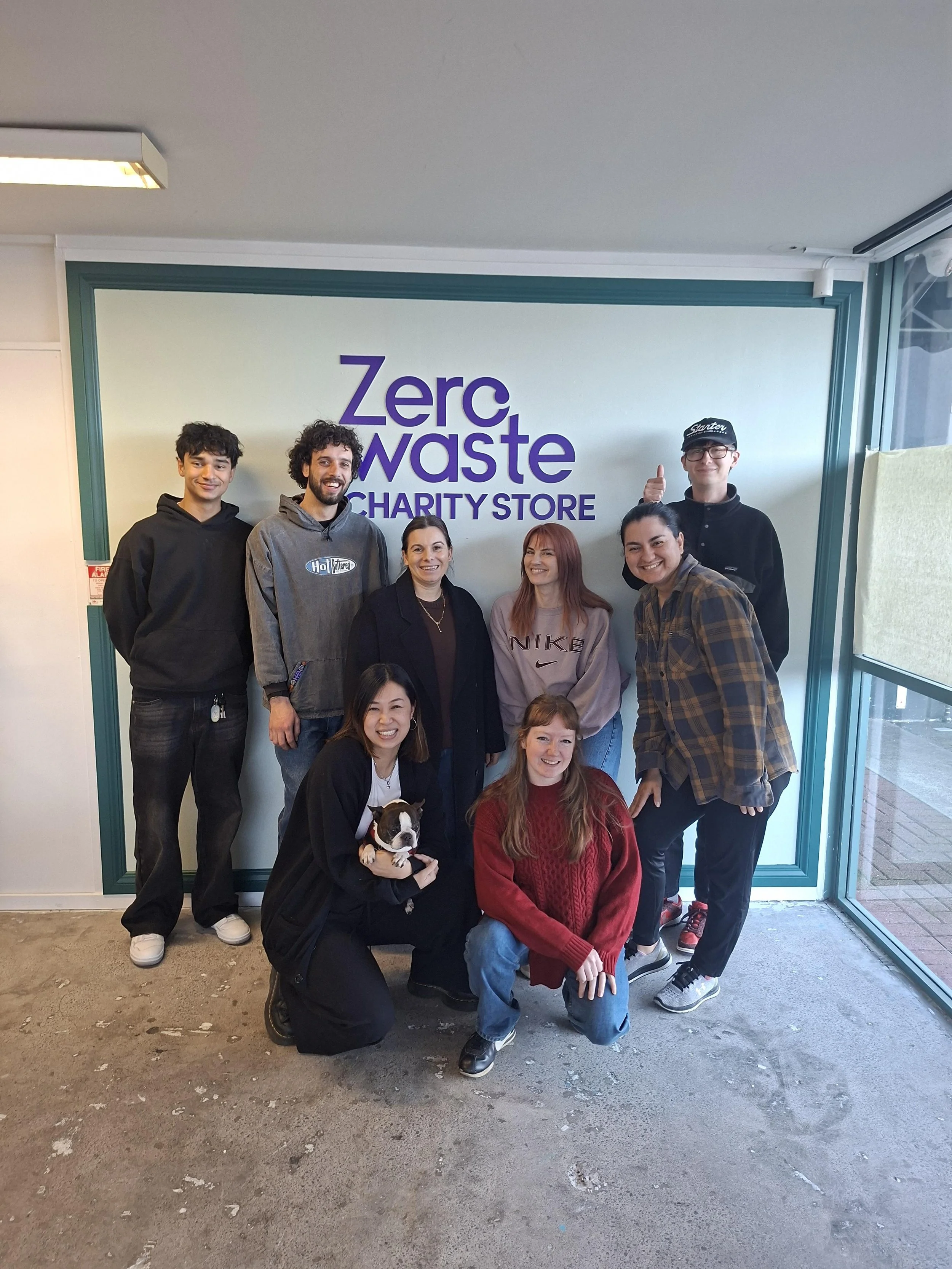 A group of nine people, including a woman holding a small dog, standing and kneeling in front of a sign that reads 'Zero Waste Chariry Store,' inside a store with glass doors and windows.