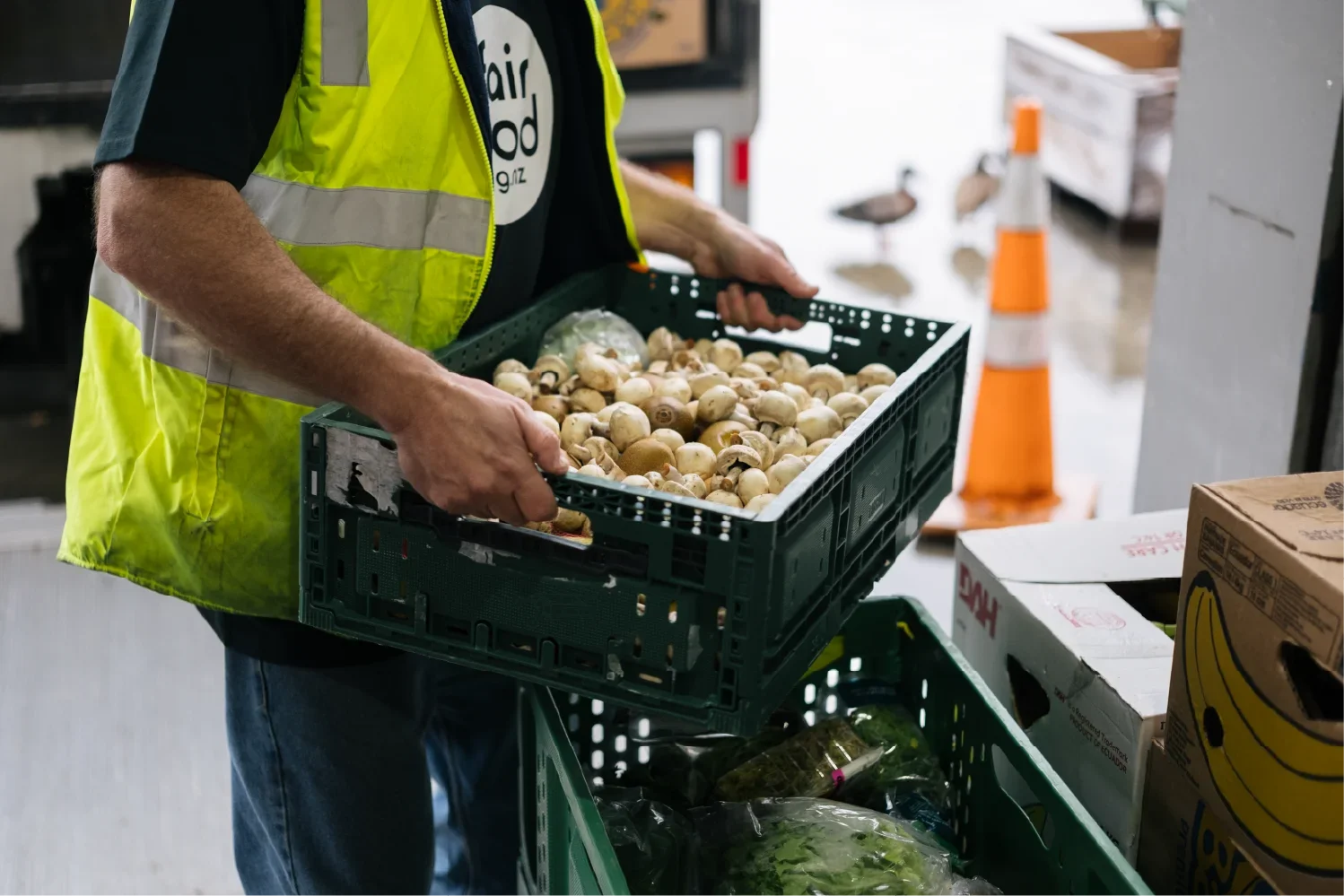 A person wearing a yellow safety vest and black shirt is holding a green plastic crate filled with button mushrooms at a warehouse. There are other produce and boxes visible in the background.