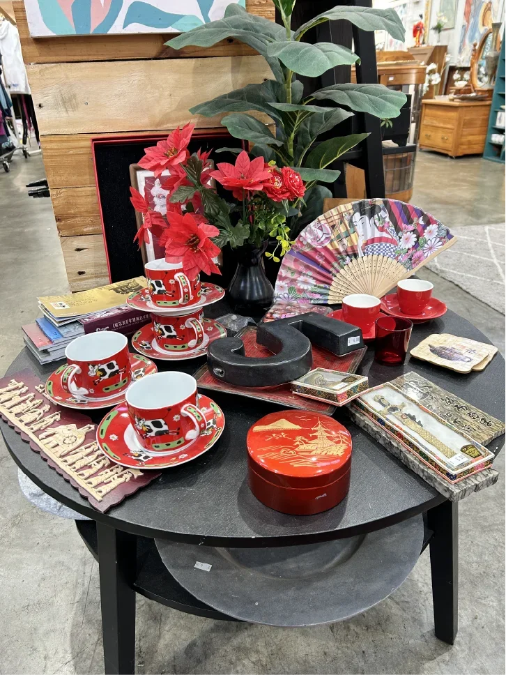 A table with various Christmas-themed decorations including red and white cups and saucers, a red round tin, a black decorative object, a small red candle holder, a decorative fan, and a vase with red poinsettia flowers and green leaves. In the background, there is a wooden wall and an indoor setting with other furniture and decor.