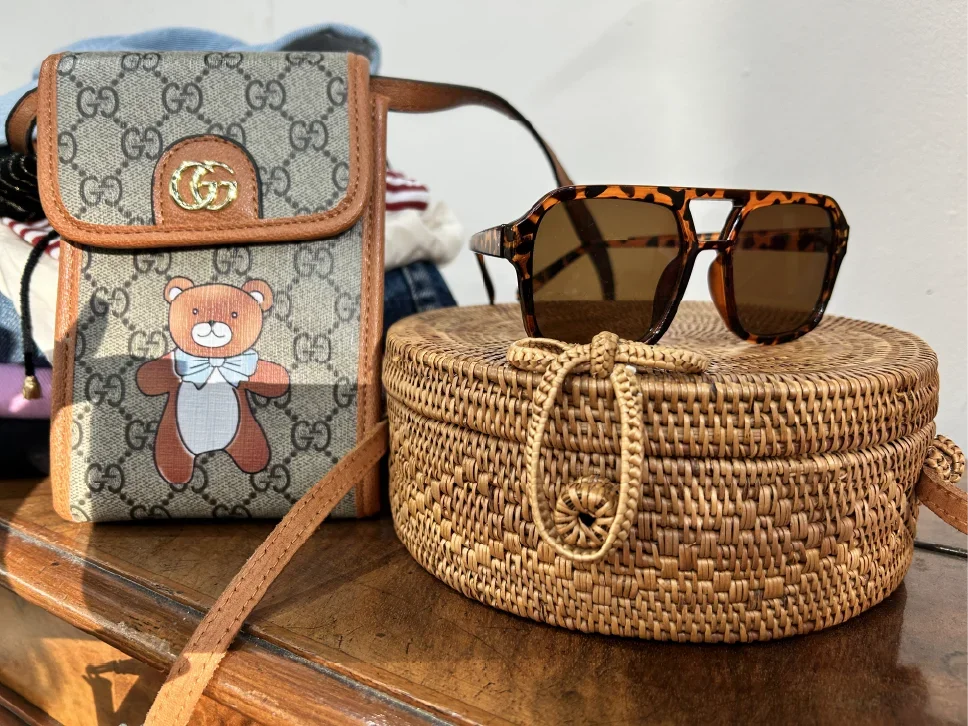 A Gucci patterned crossbody bag with a teddy bear print, a pair of tortoiseshell sunglasses on a woven basket, and a wooden surface.