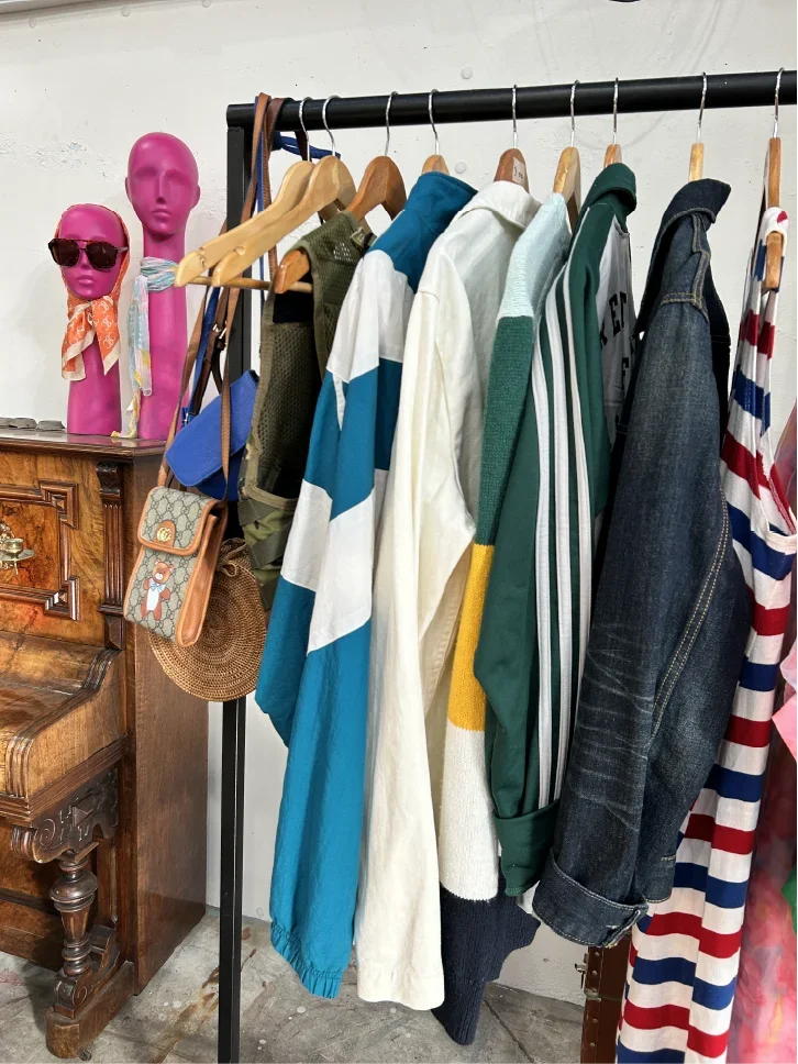 Clothes hanging on a black rack, including a blue and white striped jacket, white shirt, green and yellow jacket, dark denim jacket, and a striped dress.