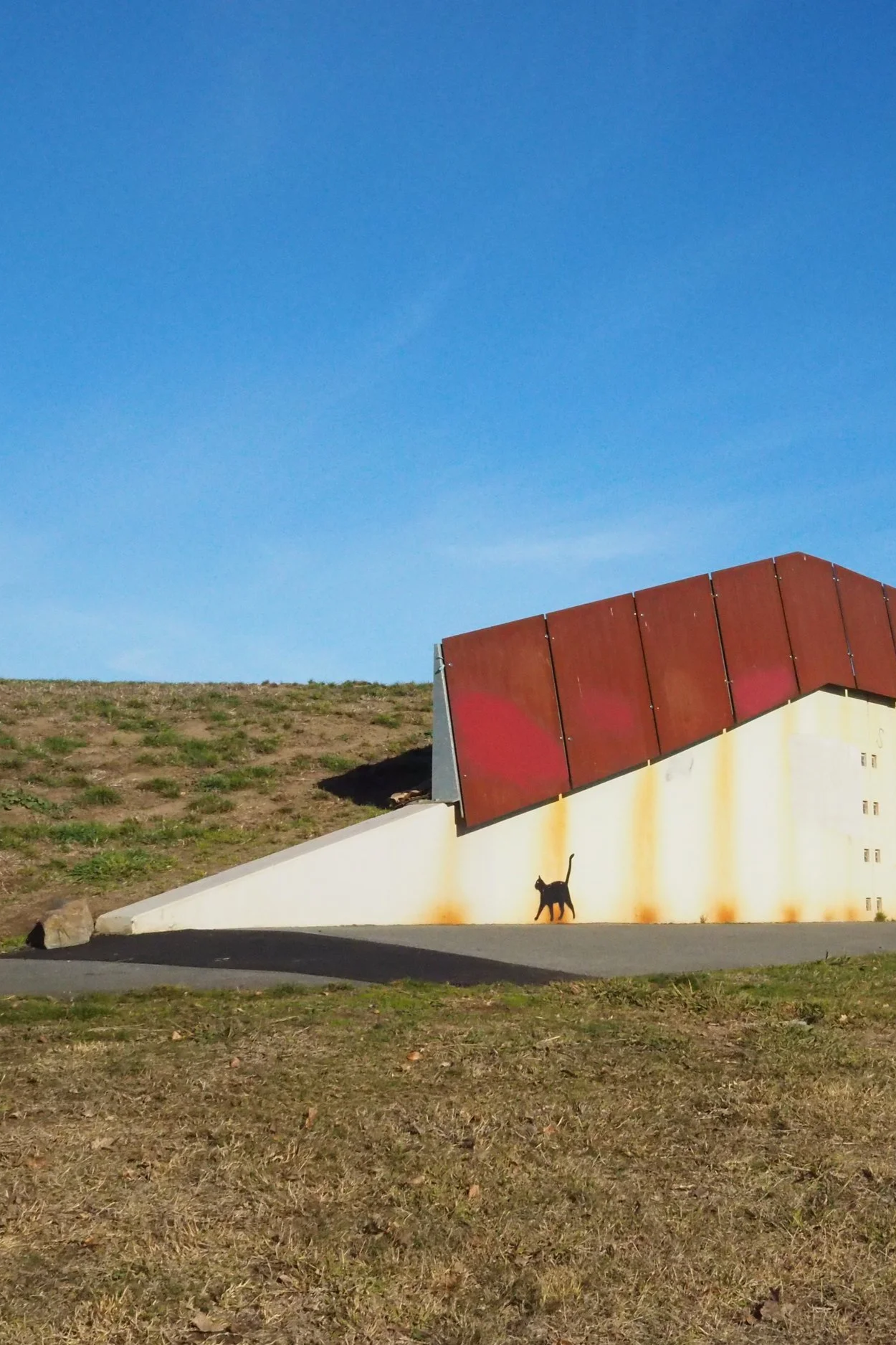 A mural on a white wall with a cat silhouette and rust-colored panels, under a clear blue sky, with a grassy hillside in the background.