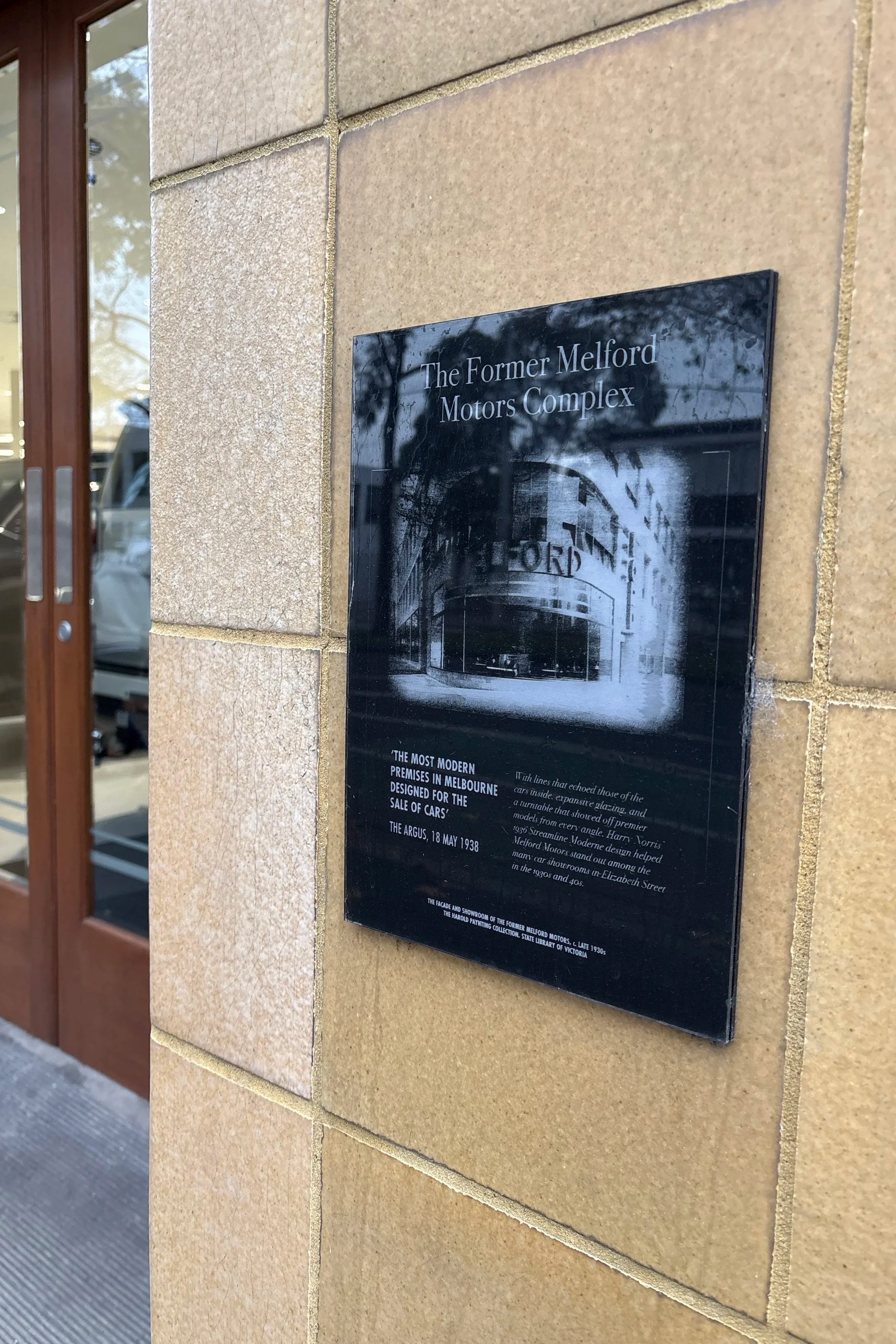 Black plaque on beige brick wall titled "The Former Melford Motors Complex" with historic information and a photograph of the complex, located outside a building with glass and wooden door.