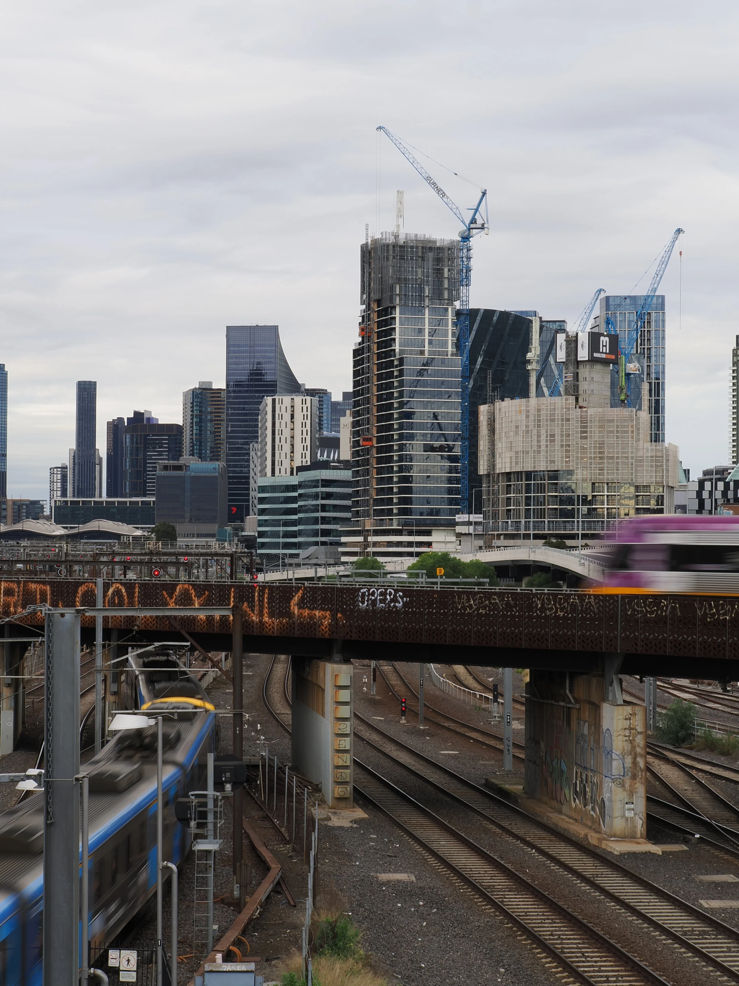 Cityscape with high-rise buildings, construction cranes on some buildings, train tracks, and a moving train in the foreground under a cloudy sky.