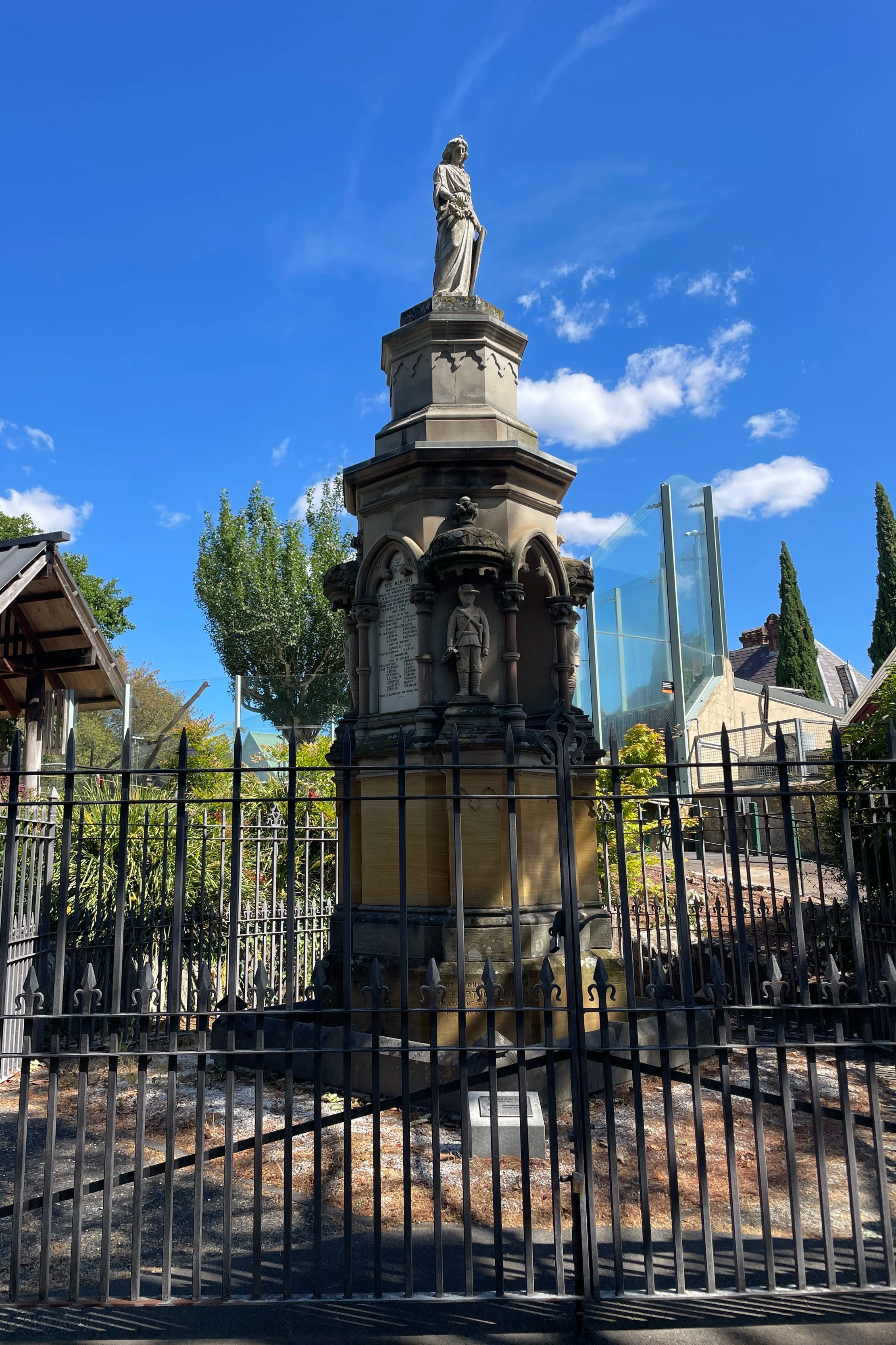 A historic monument featuring a statue of a woman atop a tall ornate pedestal enclosed by a black metal fence, with trees and modern glass structures in the background, under a clear blue sky.