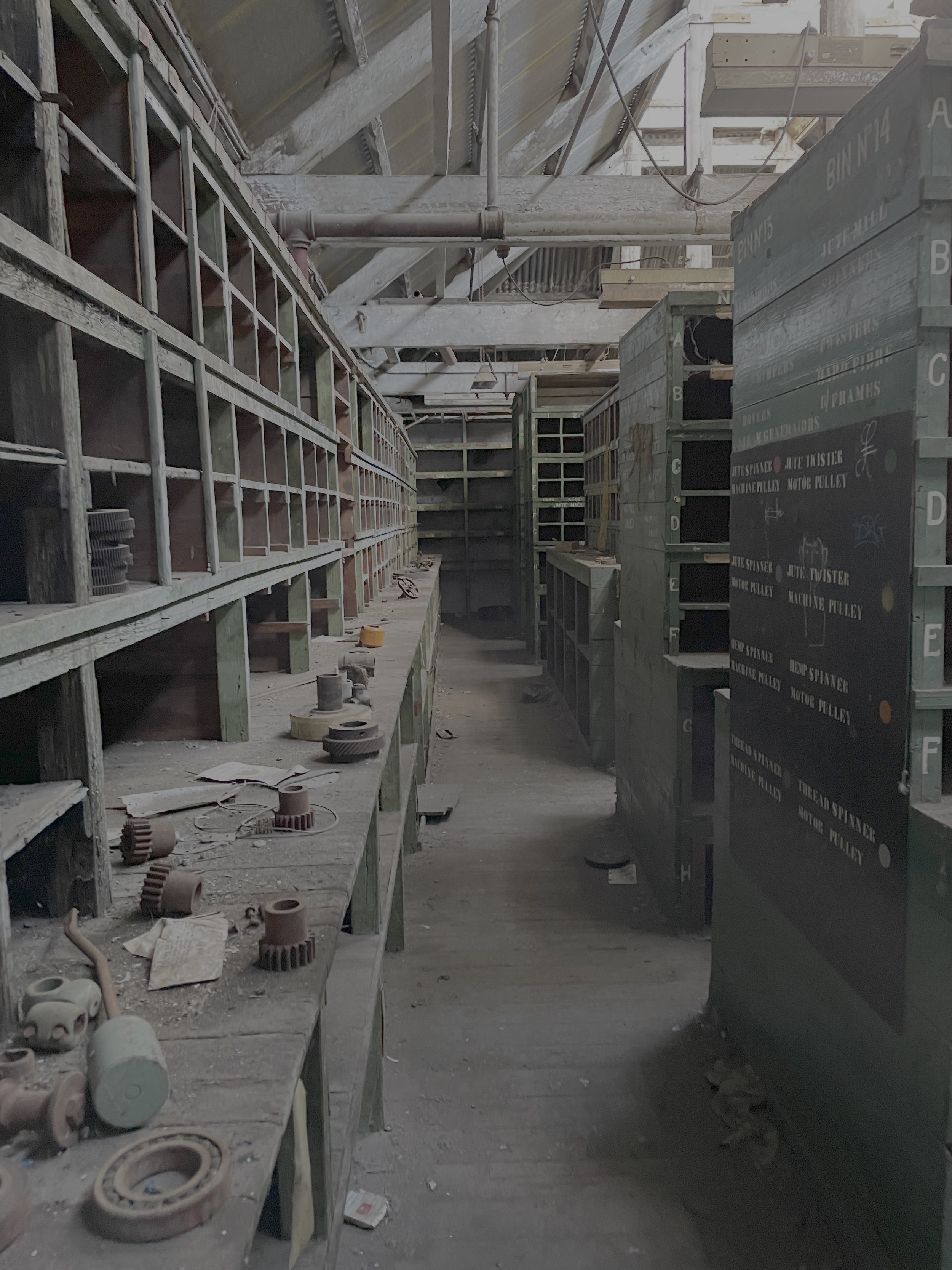 An abandoned storage room with dusty, empty shelves and scattered various small mechanical and electrical parts on the floor and shelves.