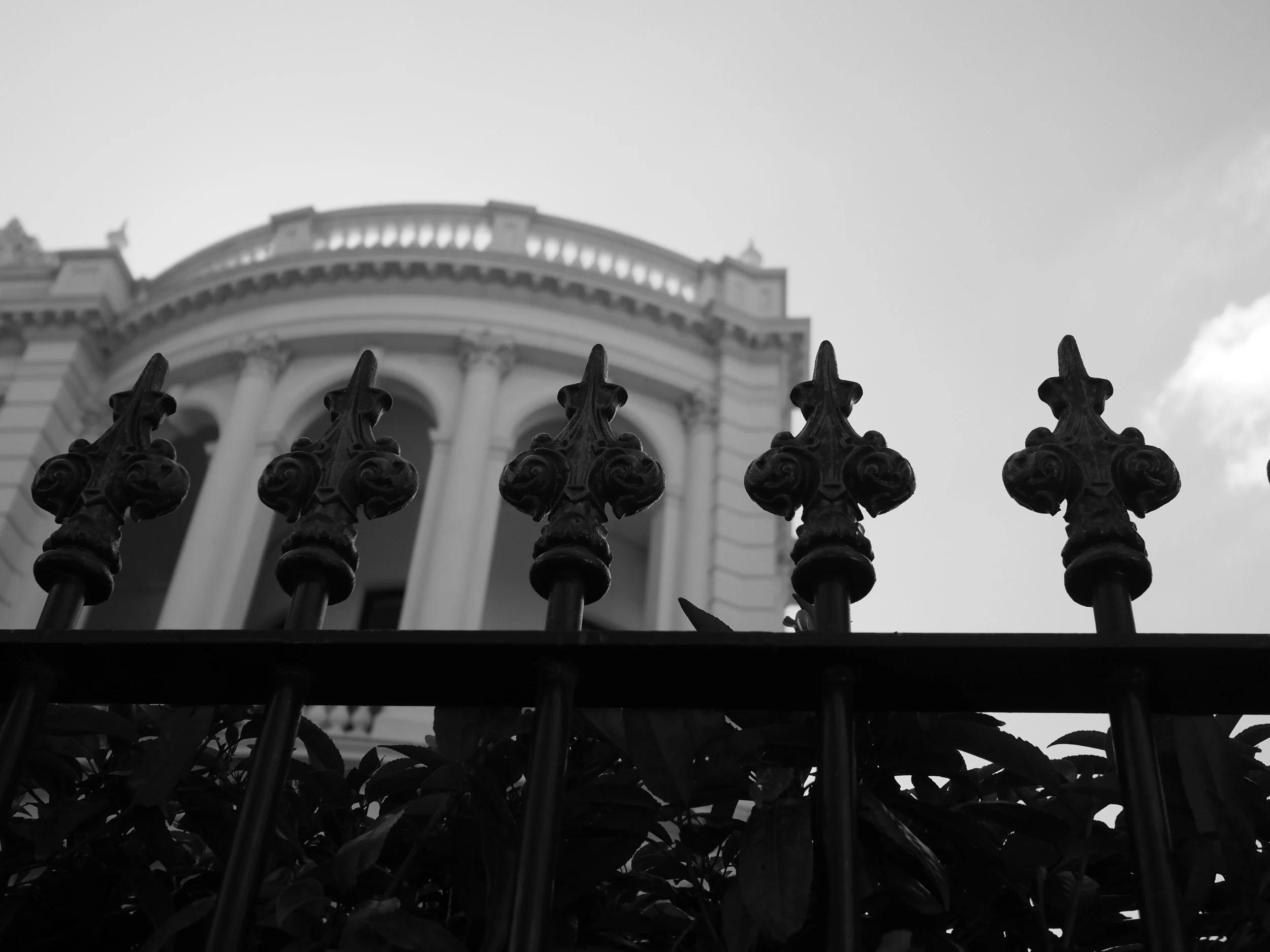 View of a historic building with classical architecture behind a decorative iron fence, taken from below with the sky in the background.