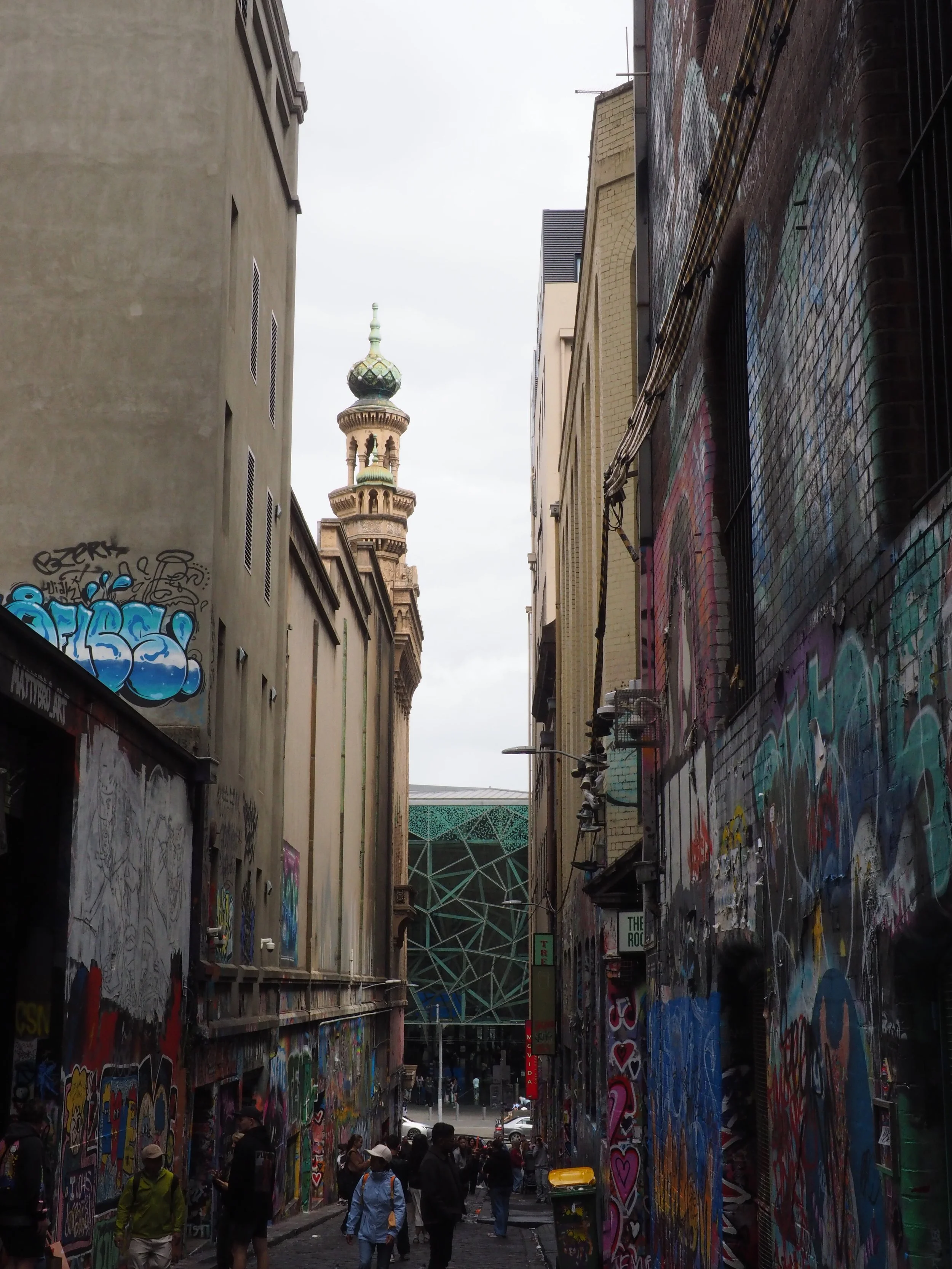A bustling city alleyway with graffiti-covered walls, crowds of people, and an architectural landmark in the background.