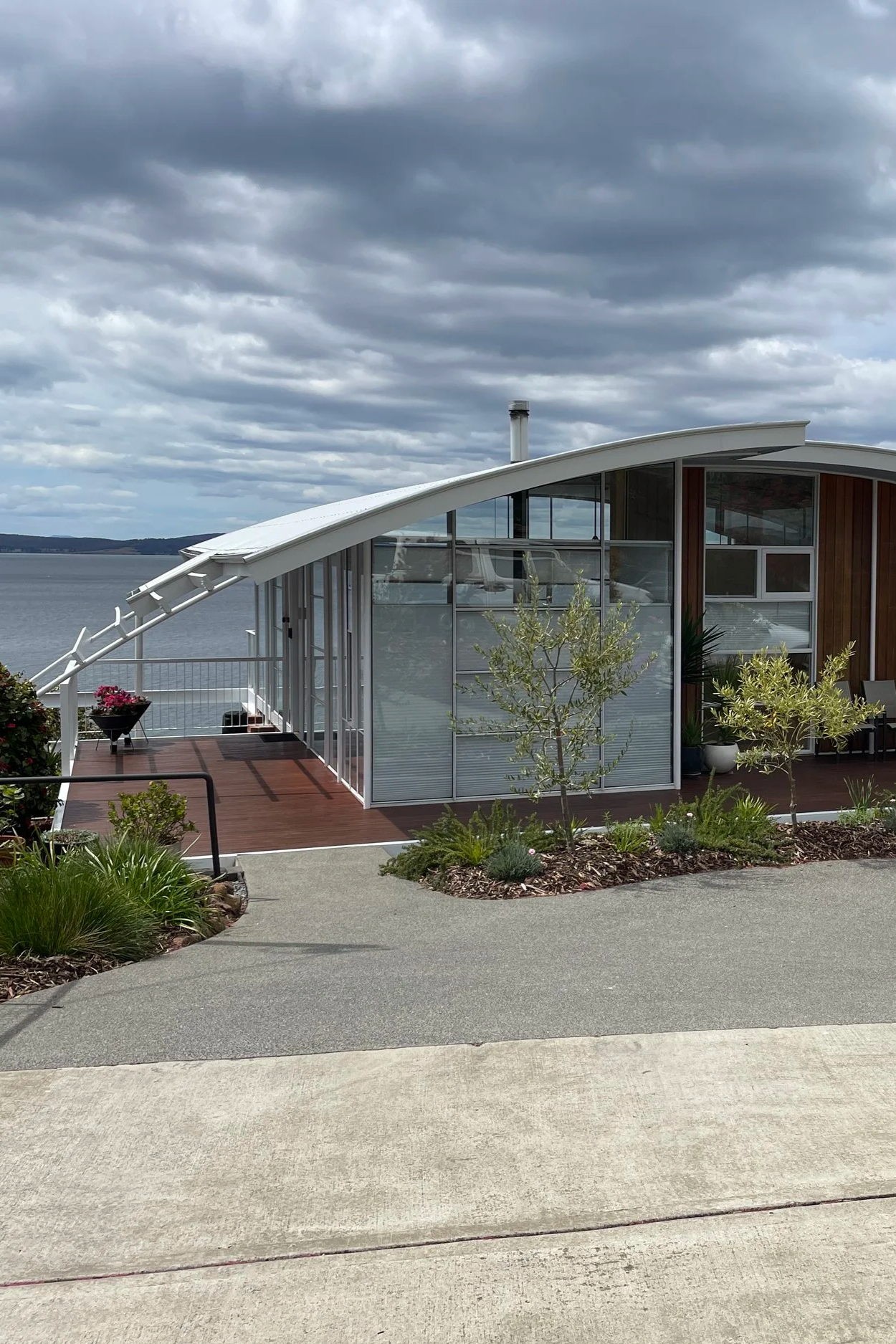 A modern house with large glass windows and a curved roof overlooking a body of water under cloudy skies, surrounded by small trees and landscaped garden.