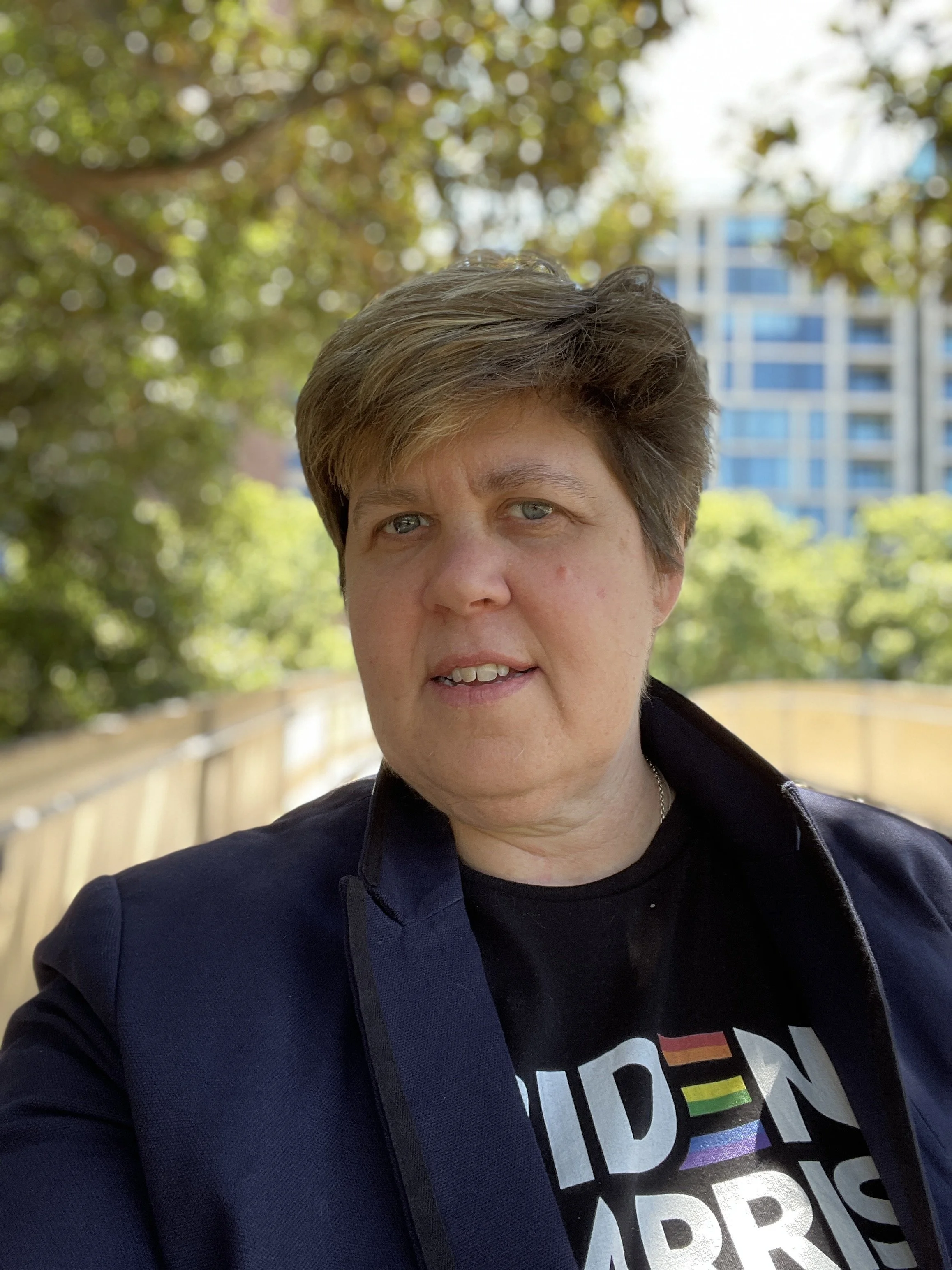 Close-up of Dr Meighen Katz face, wearing a black jacket and a T-shirt with a rainbow-colored pride flag and wordings. Background shows green tree leaves and a building.