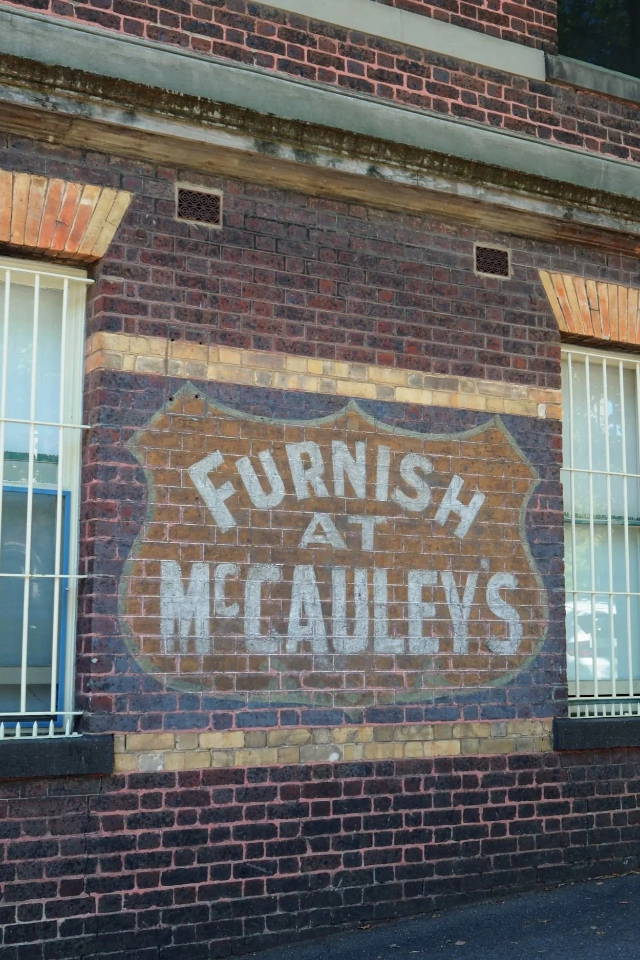 A brick building with a painted sign on the wall. The wall has two windows with white bars and a decorative brick arch above each window. The sign appears faded and worn.