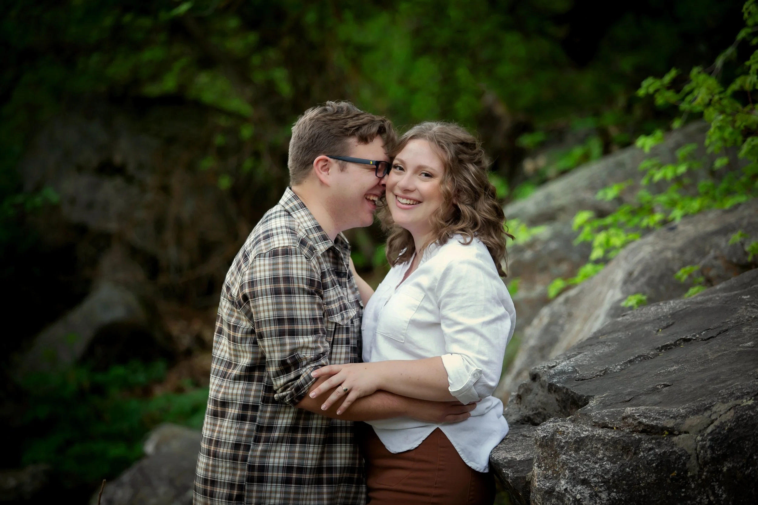 A couple is smiling and embracing outdoors, surrounded by greenery and rocks.