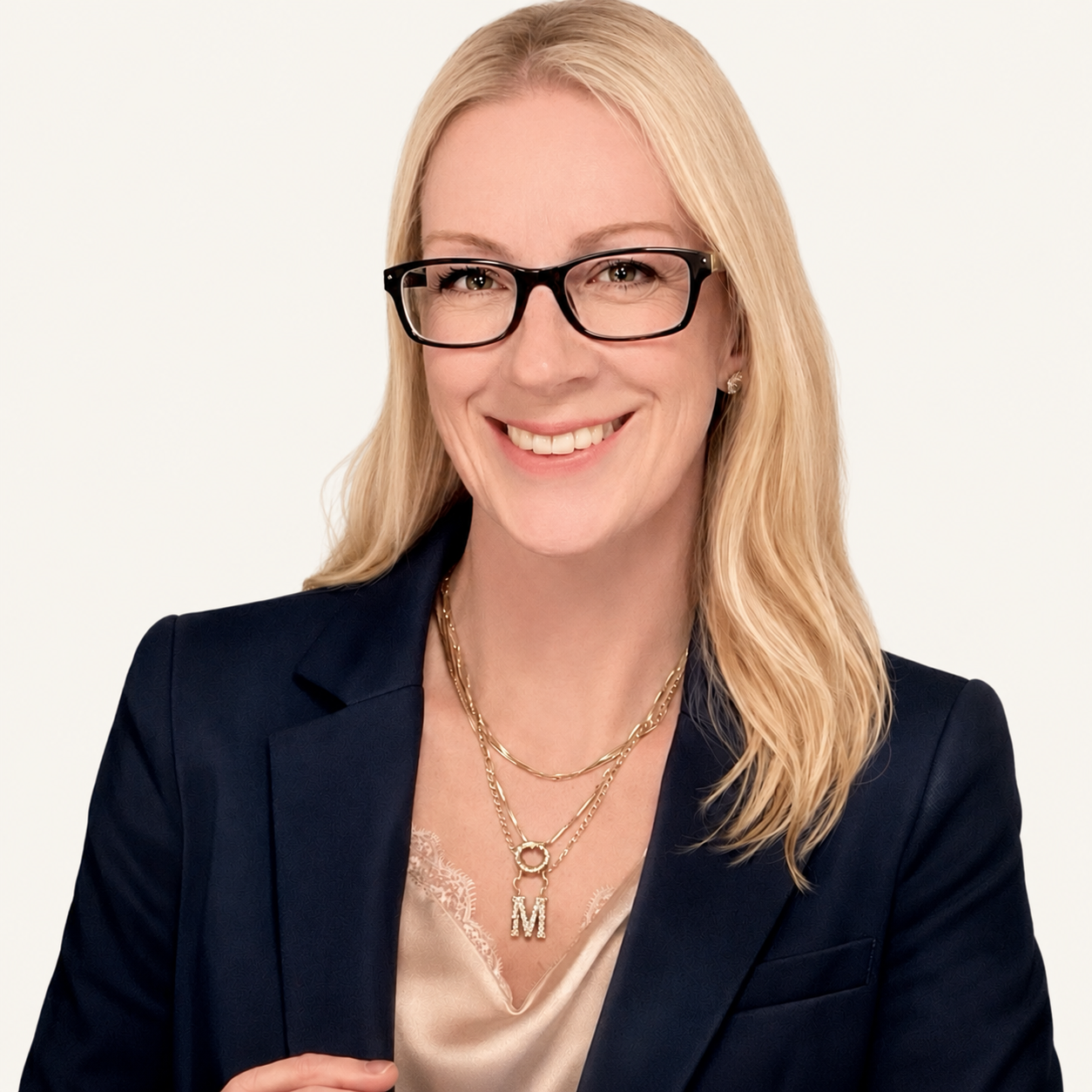 A smiling woman with blonde hair, black glasses, wearing a dark blazer, pearl earrings, and layered gold necklaces with a monogram 'M' in the center.
