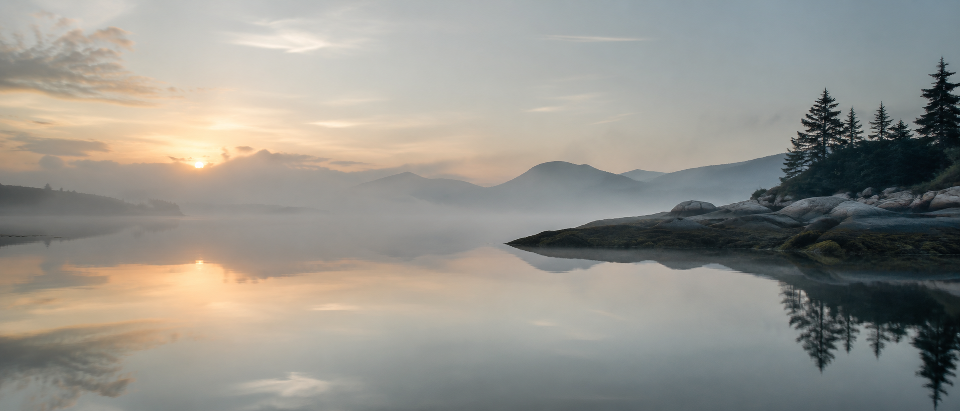 A serene landscape of a calm lake at dawn, surrounded by mist-covered mountains and pine trees, with the sun low on the horizon and clouds reflected in the water.