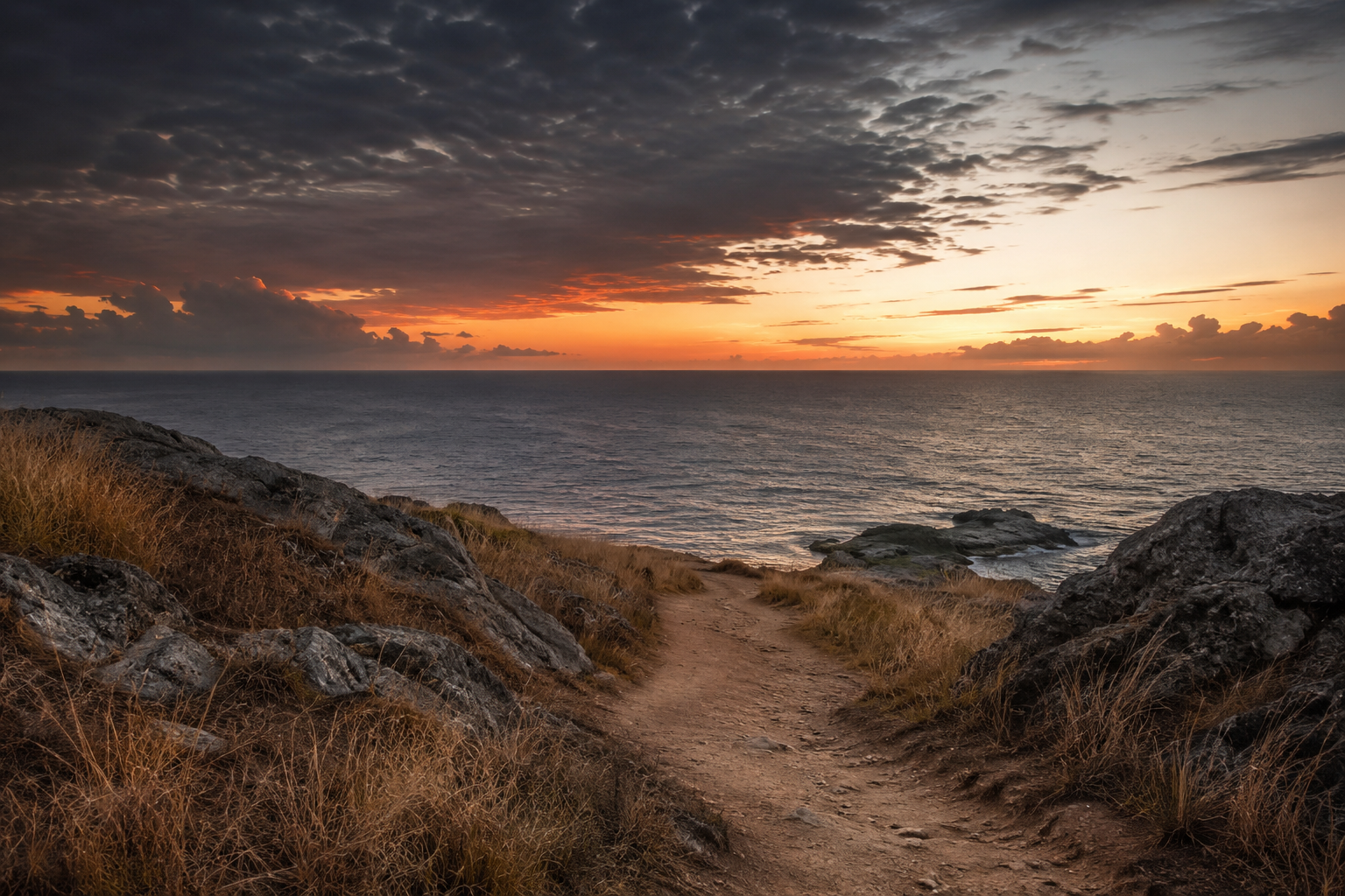 A dirt trail leading towards the ocean during sunset, with rocky and grassy terrain on either side, and a mostly cloudy sky with some orange and yellow hues.
