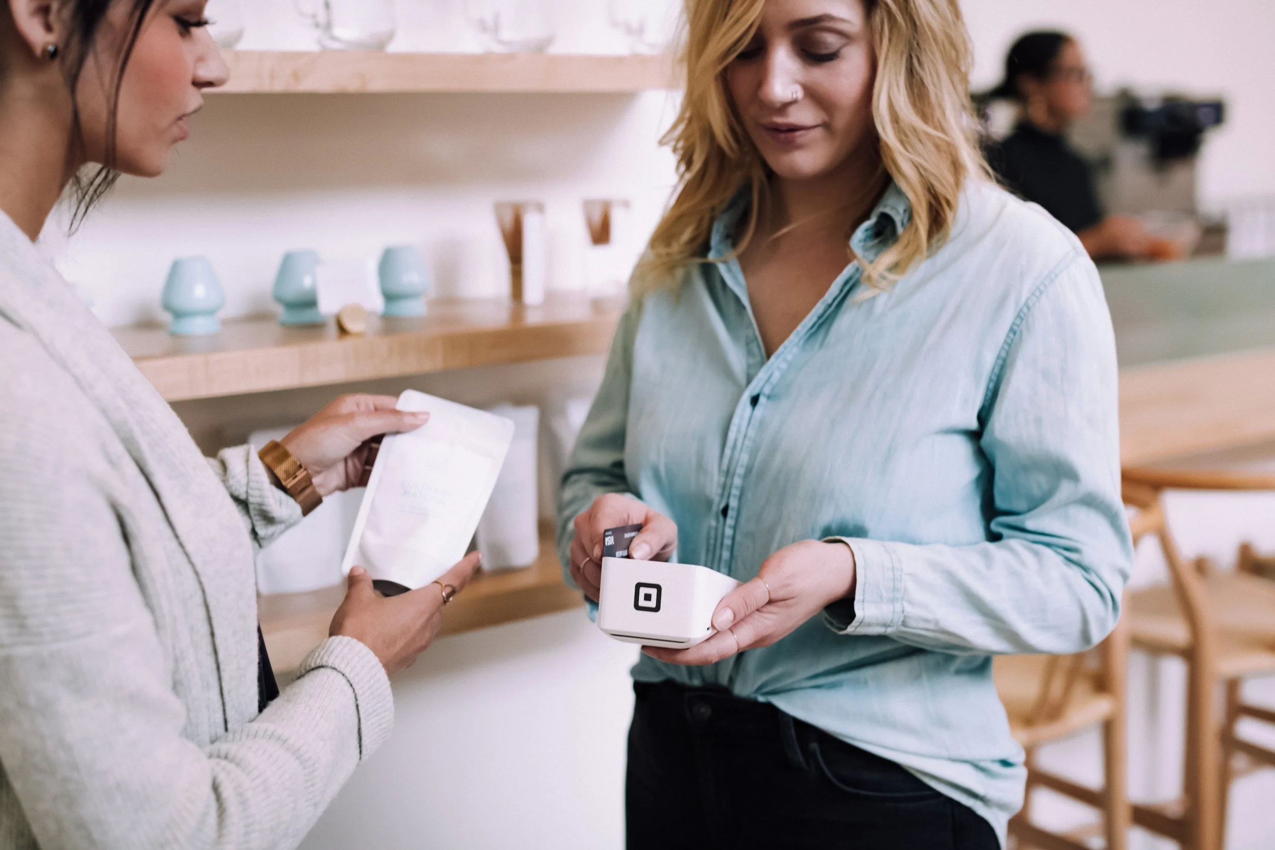 Two women exchange a payment in a retail store, one woman hands over a black card while the other holds a white device with a QR code. Book Keeping