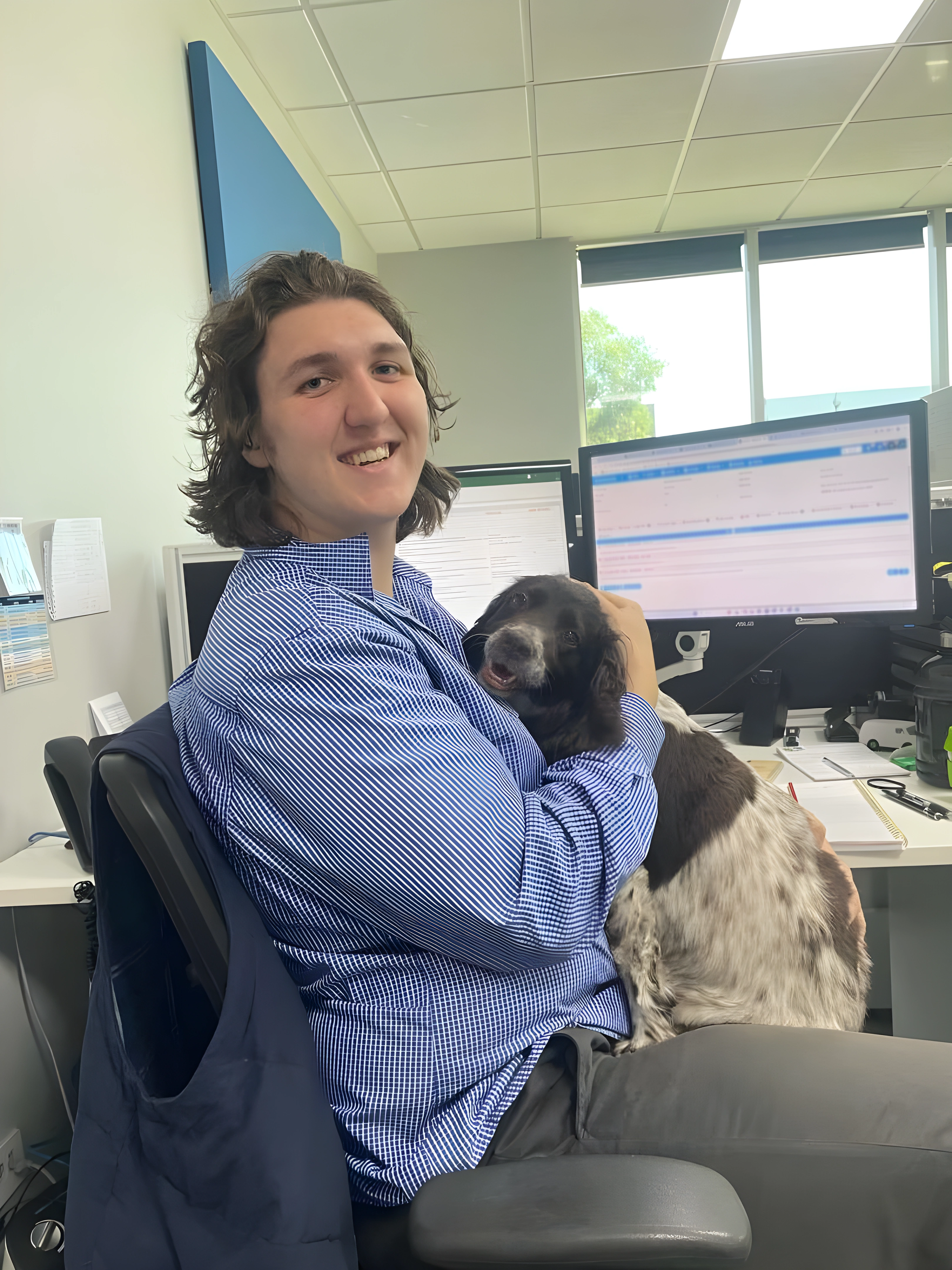 Person with shoulder-length hair sitting at a desk in front of a computer screen, holding a dog on their lap, in an office setting with windows and office supplies visible. Book Keeping