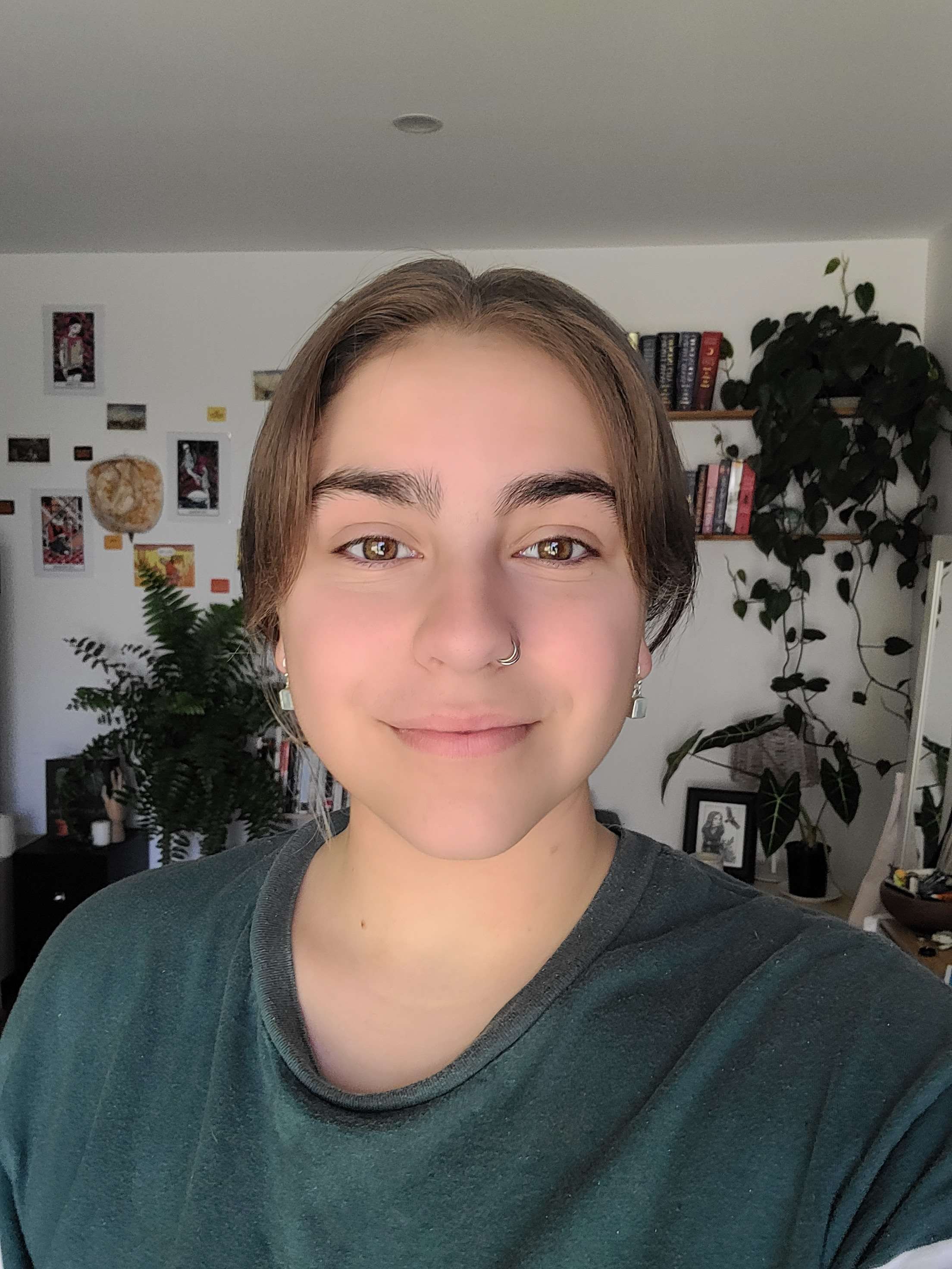 A young person with short brown hair, hazel eyes, and earrings, smiling in a room with plants, bookshelves, and wall decorations. Book Keeping