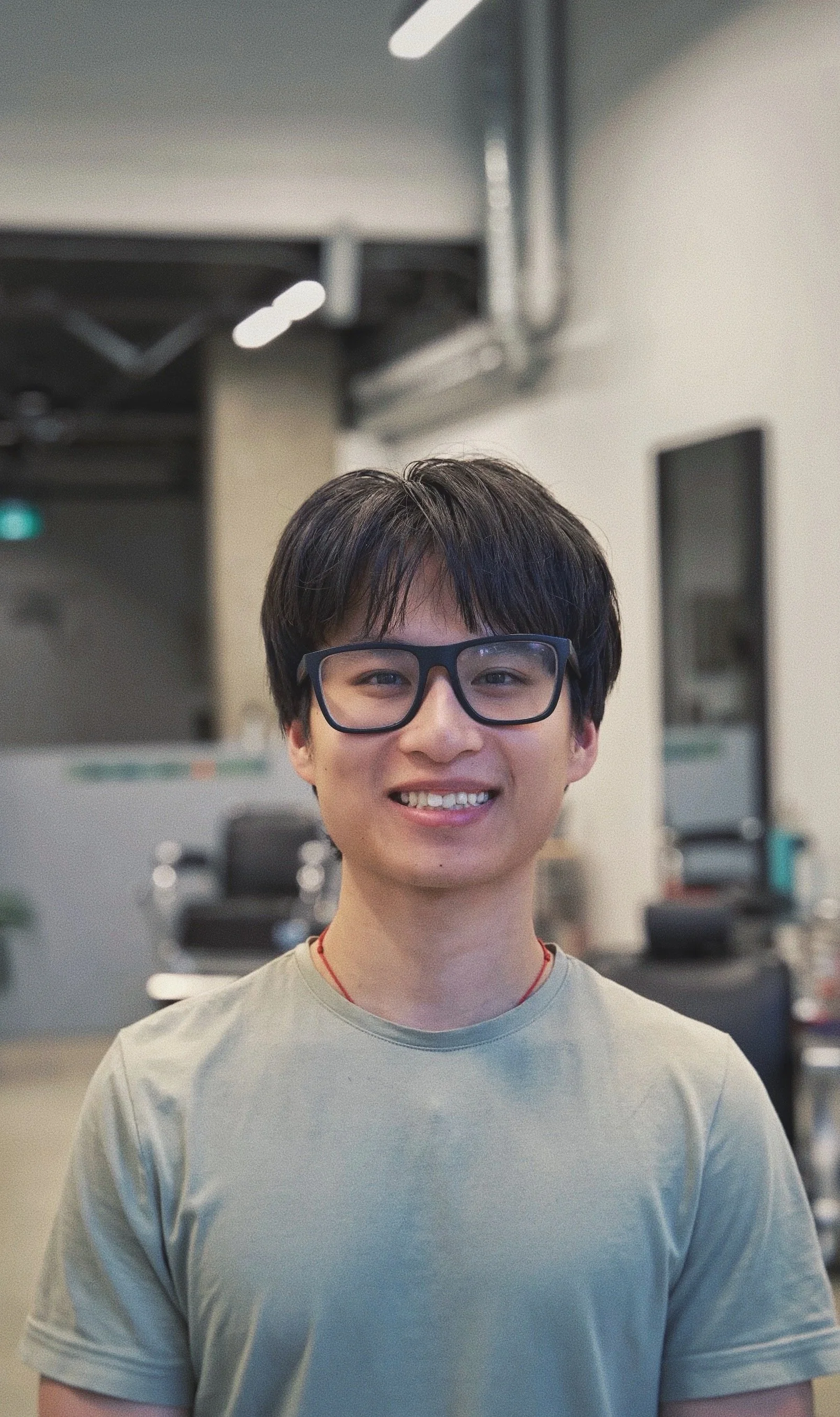 A young man with short dark hair, wearing glasses and a beige t-shirt, smiling in a gym or fitness center.
