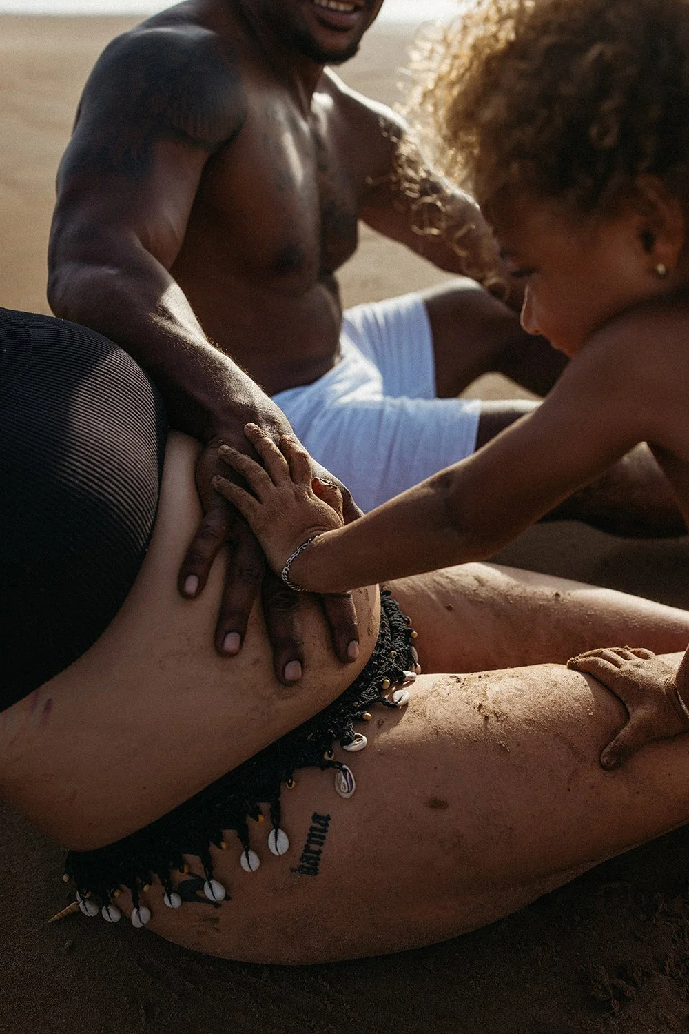 A family photo of a pregnant woman, her toddler and partner's hands on her belly on the beach in Darwin by Captured by Cody Photography.
